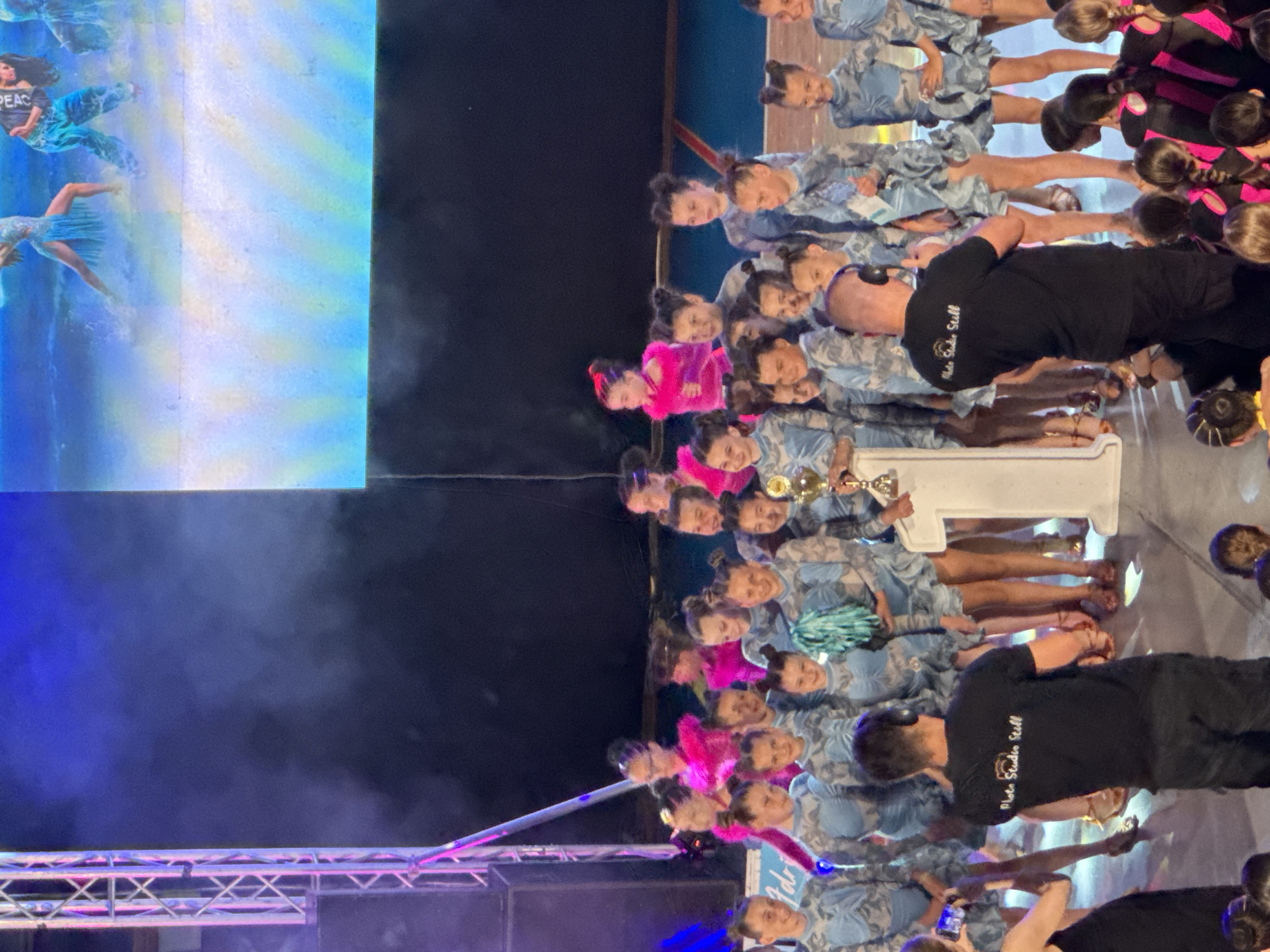 The image depicts a group of young dancers on a stage in Bar, Montenegro. The dancers are predominantly girls, dressed in matching blue and grey outfits with ruffled skirts. Some are holding a trophy and a turquoise pom-pom. A man in a black t-shirt with "Photo Studio Still" printed on the back is standing in the foreground, facing away from the camera. A large screen behind the dancers displays a blue and white image. The stage is lit with stage lighting, and there is some smoke or fog effect. The overall scene suggests an awards ceremony or performance event.