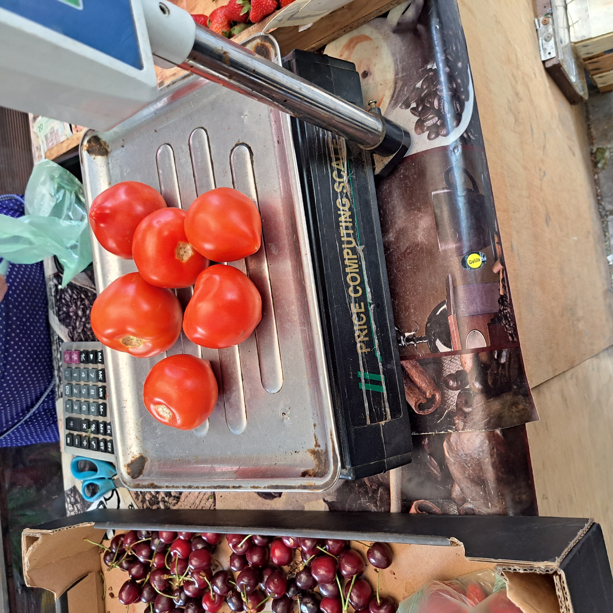 This image captures a close-up view of a fruit and vegetable stall, likely located in Odesa, Ukraine, as indicated by the context. The foreground is dominated by a metal scale, on which six bright red tomatoes are neatly arranged. Below the scale, a black base is visible, displaying the faded text "PRICE COMPUTING SCALE" with some tape applied over it. The scale itself rests on a surface adorned with a printed image of coffee beans and a coffee grinder. To the left of the scale, a cardboard box overflows with a generous pile of dark red cherries, their stems still attached. A pair of bright blue scissors is also visible near the cherries, next to a calculator. In the background, out of focus, a person wearing a blue and white patterned garment is partially visible, and a green plastic bag is also present. The overall scene suggests a market setting, where fresh produce is being weighed and prepared for sale.