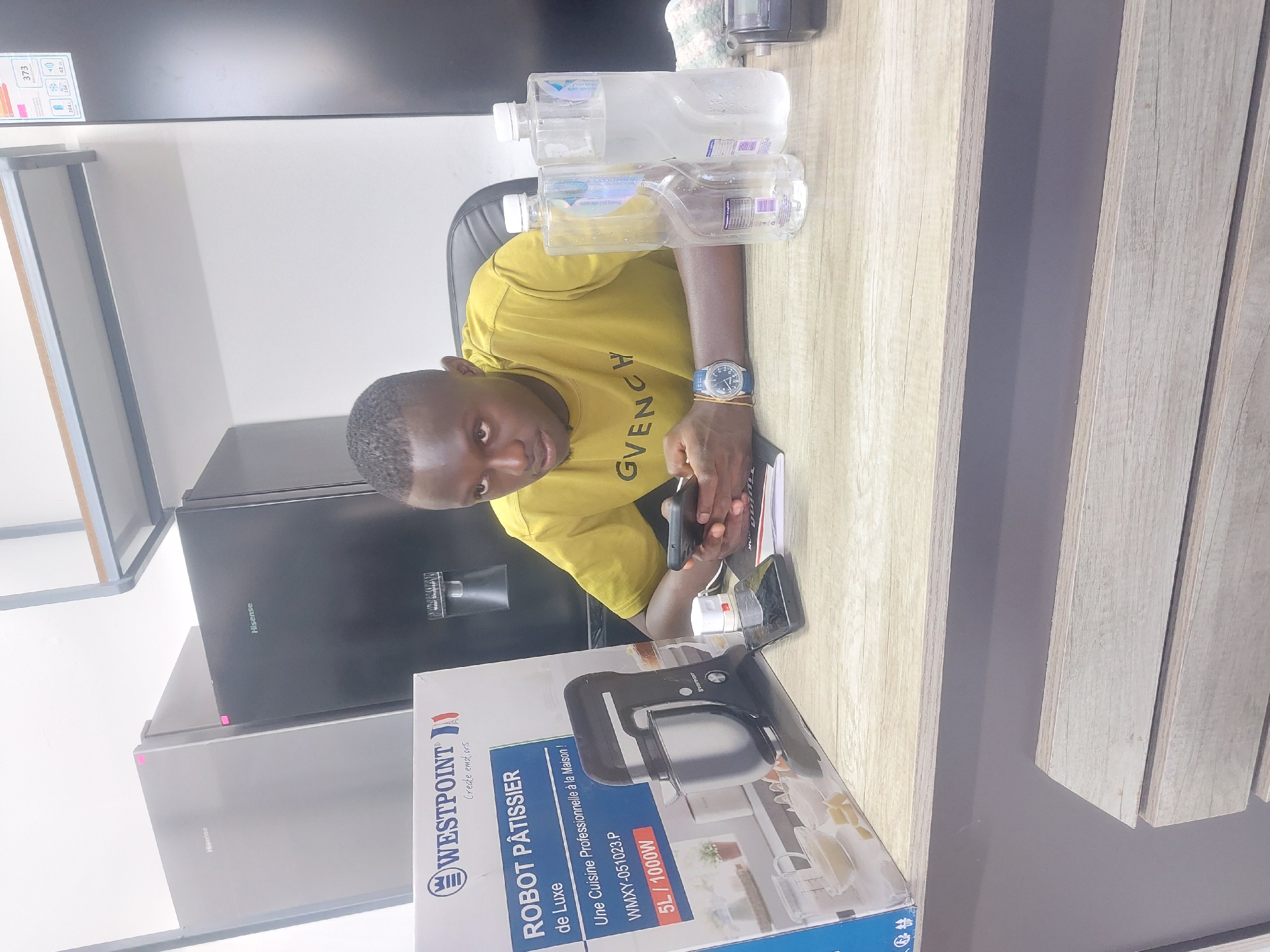 The image depicts a man seated at a desk. He is dark-skinned, wearing a yellow t-shirt with "GVENCHY" printed on it, and a blue watch. He is holding a smartphone and looking at the camera.
On the desk, there are two water bottles, a Westpoint Robot Pâtissier de Luxe box, a notebook, and another smartphone. The Westpoint box indicates it's a 5L/1000W food processor. Behind the desk, there is a black Hisense refrigerator. There appears to be a price tag on the top of the refrigerator on the left. The room is indoors and appears to be a shop or showroom, judging by the presence of appliances.
Based on the language on the Westpoint box and the presence of Hisense appliances, it is plausible this image was taken in Tanzania, particularly in Dar es-Salaam, as Westpoint products are sold there.