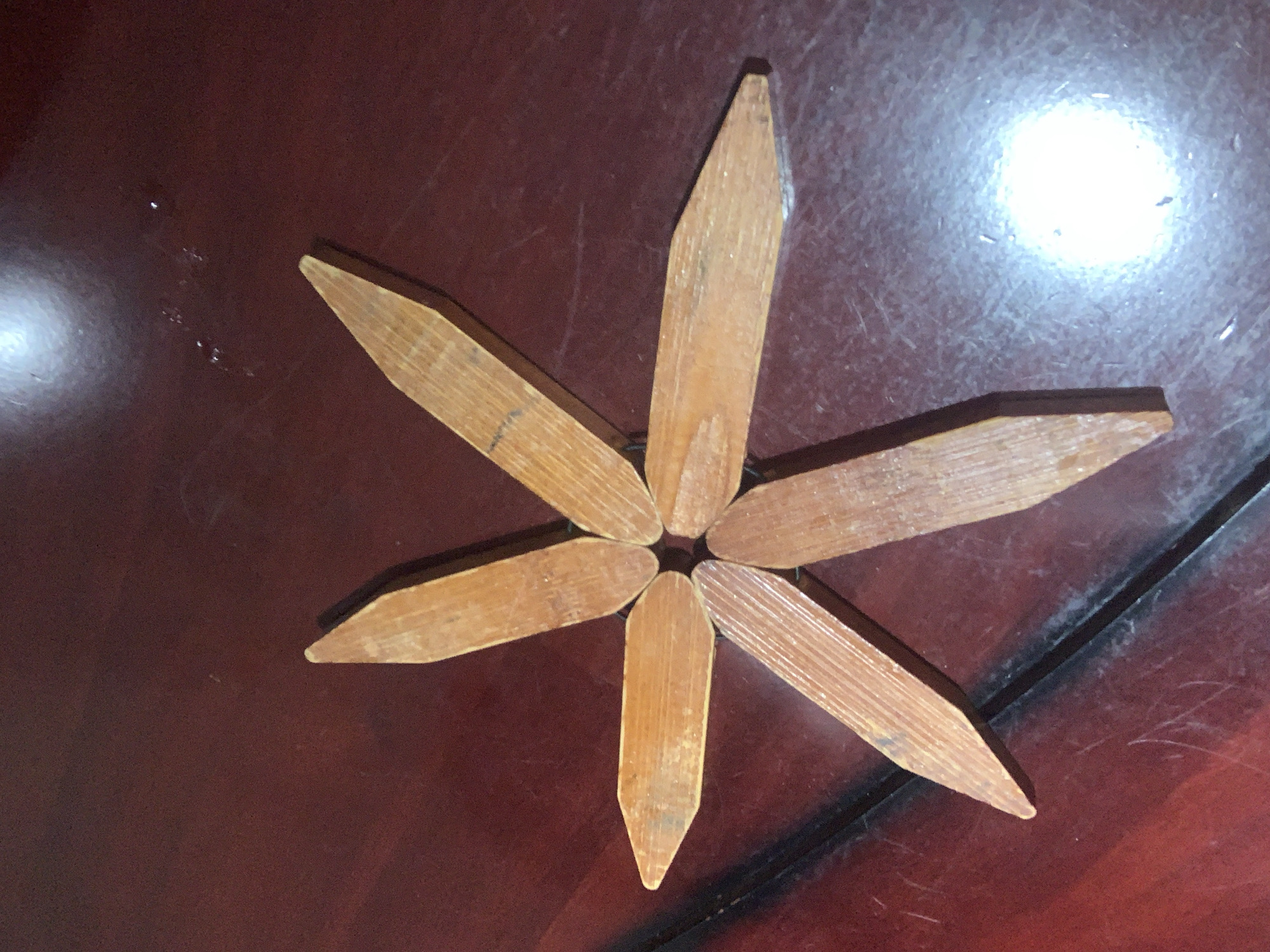 A six-pointed star shape, composed of wooden pieces with pointed ends, is arranged on a dark, polished wooden surface. The star is approximately centered in the frame. Each wooden piece is approximately 6 inches long and tapers to a point. The wood appears aged and shows visible grain patterns. The surface beneath the star has a slight sheen and exhibits a few light scratches and smudges, suggesting it is a table or countertop. A bright, circular light reflection is visible in the lower right quadrant of the image, indicating an overhead light source. No people or animals are present in the image. The scene is indoors. The precise location within Abuja, Nigeria cannot be determined from the image alone.
