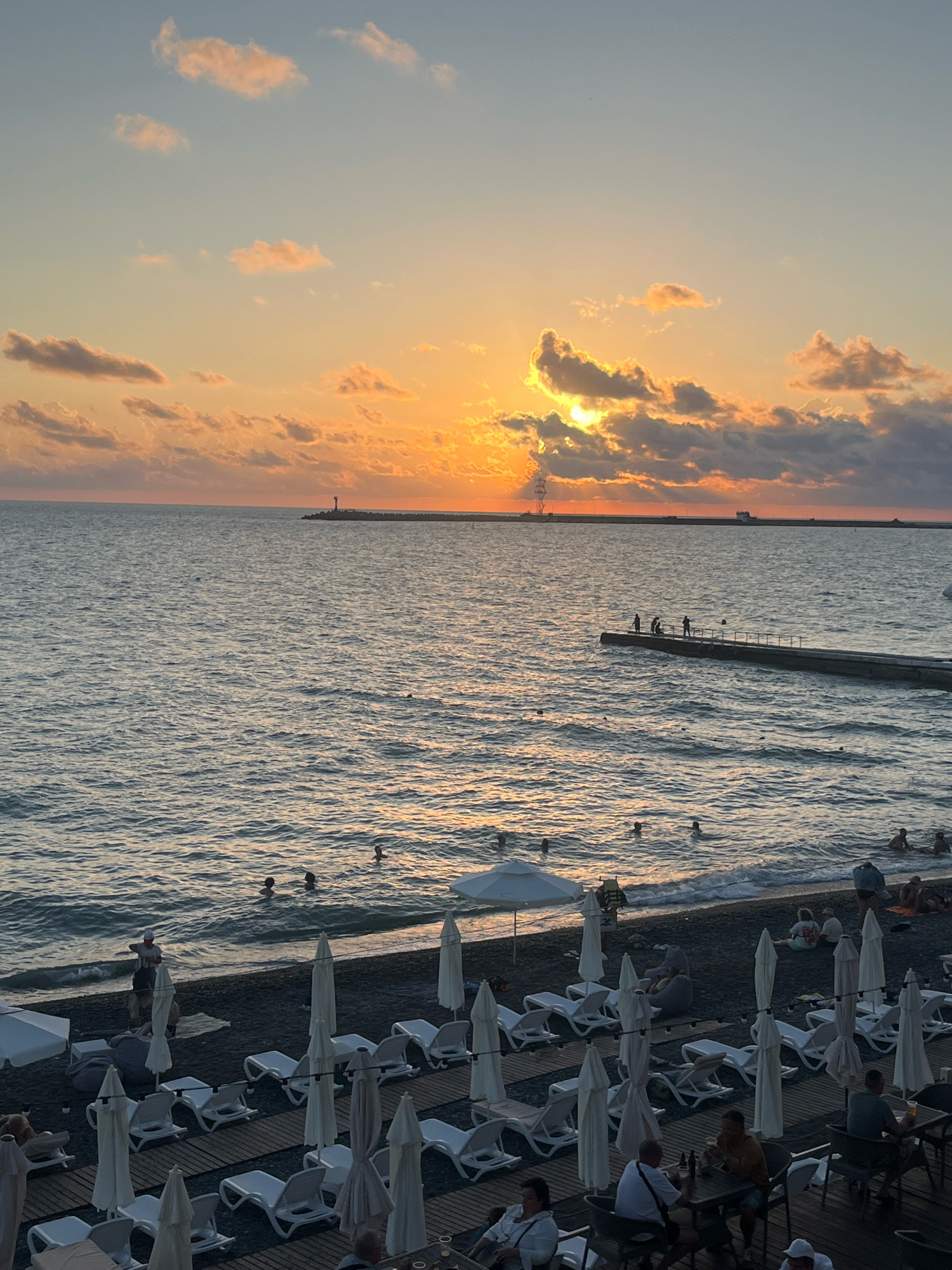 This is an eye-level, vertical, medium shot of a beach scene at sunset in Sochi, Russia. The sun is setting over the Black Sea, casting a warm orange and yellow glow across the sky and reflecting on the water's surface. The clouds are a mix of pink, orange, and gray. In the distance, a breakwater with a lighthouse and industrial structures extends into the sea.
Closer to the shore, several people are swimming or wading in the water. A pier extends from the right side of the image, with a few individuals standing on it. The beach itself is dark-colored, with rows of white lounge chairs and closed umbrellas set up on wooden decking. More people are seated at tables on a patio area in the foreground, some of whom are holding drinks. A person is standing near the water's edge, possibly walking along the shore. The overall atmosphere is one of a relaxing evening at the beach.