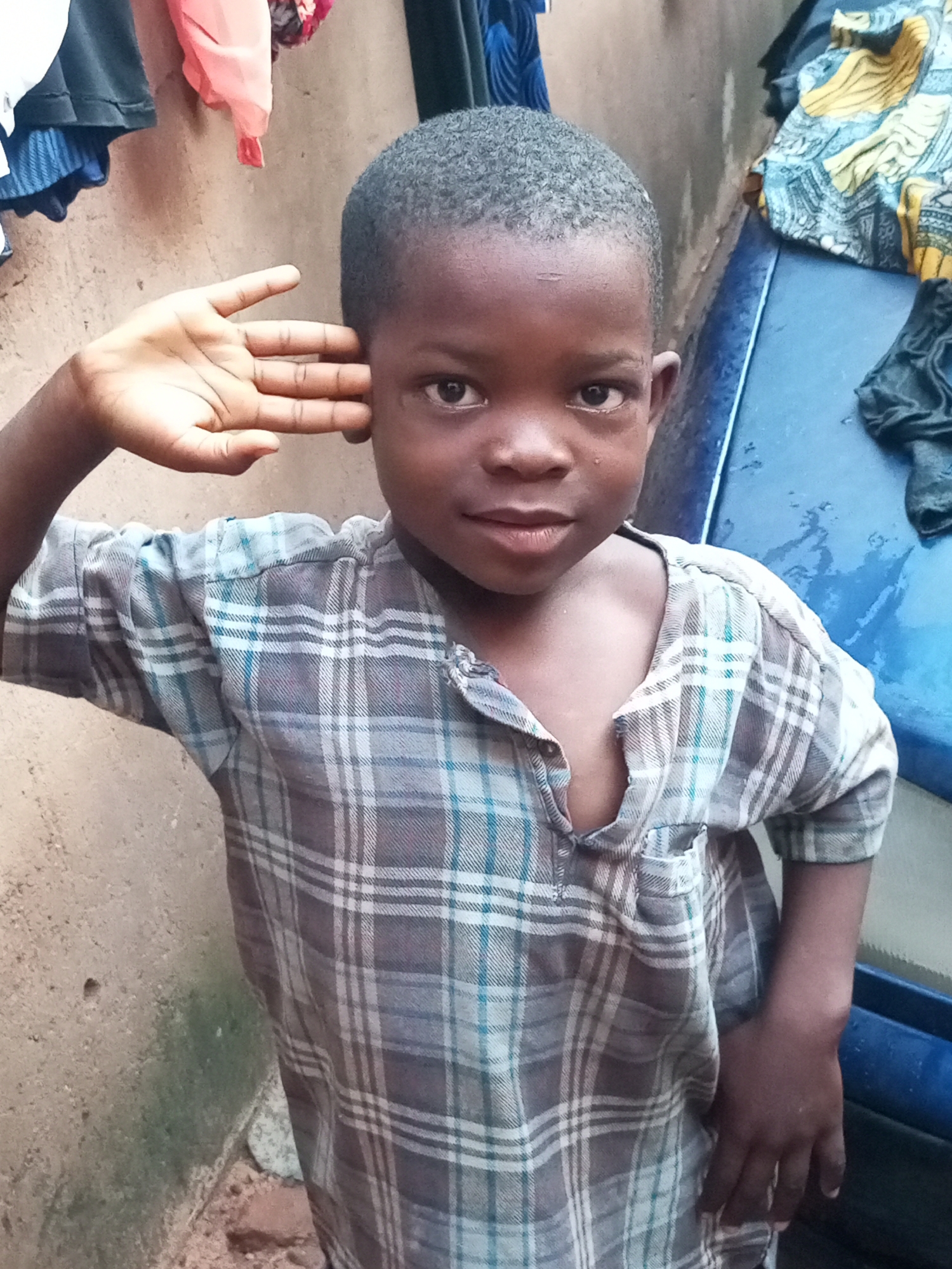 A young Black child, likely a boy, is the sole subject of this medium shot photograph. He is wearing a plaid shirt with a collar and a buttoned placket, in shades of grey, white, and light blue. His right hand is raised to his head, with fingers gently touching his temple and ear. His gaze is directed towards the camera with a neutral, perhaps slightly curious, expression.
The background is divided into two main areas. To the left, a rough, textured wall, likely made of concrete or stucco, has a slightly grimy appearance with some green patches that could be moss or algae. Above the wall, various items of clothing are hanging, including a pink garment, a black t-shirt, and a blue patterned fabric. To the right, a vibrant blue surface, possibly a vehicle or a large plastic container, occupies the space. This surface appears wet, with visible water droplets and streaks. A patterned fabric with yellow and blue designs is draped over this blue surface, and a black garment is also visible nearby.
The lighting suggests it is daytime, but the overall atmosphere is somewhat subdued, possibly due to overcast conditions or the presence of shade. The rough textures of the wall and the casual arrangement of the clothing hint at an outdoor or semi-outdoor setting in a somewhat utilitarian or informal environment. The presence of water on the blue surface could indicate recent rain or a washing activity. No visible text is present in the image. The setting in Tempure, Nigeria, adds a layer of geographic context to the scene.