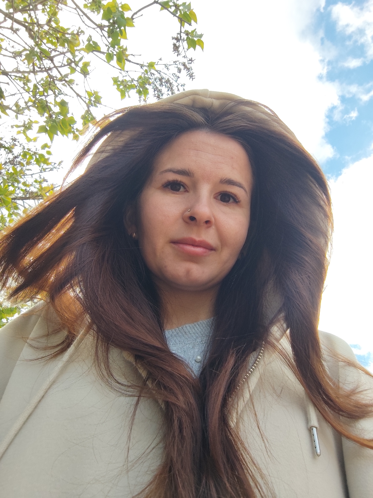 A woman with long, brown hair is taking a selfie outside. She is wearing a light-colored hooded jacket and a light blue sweater. Part of a tree with green leaves is visible in the background, along with a blue sky with scattered clouds. There is no indication the location is Фоки, Russia.
