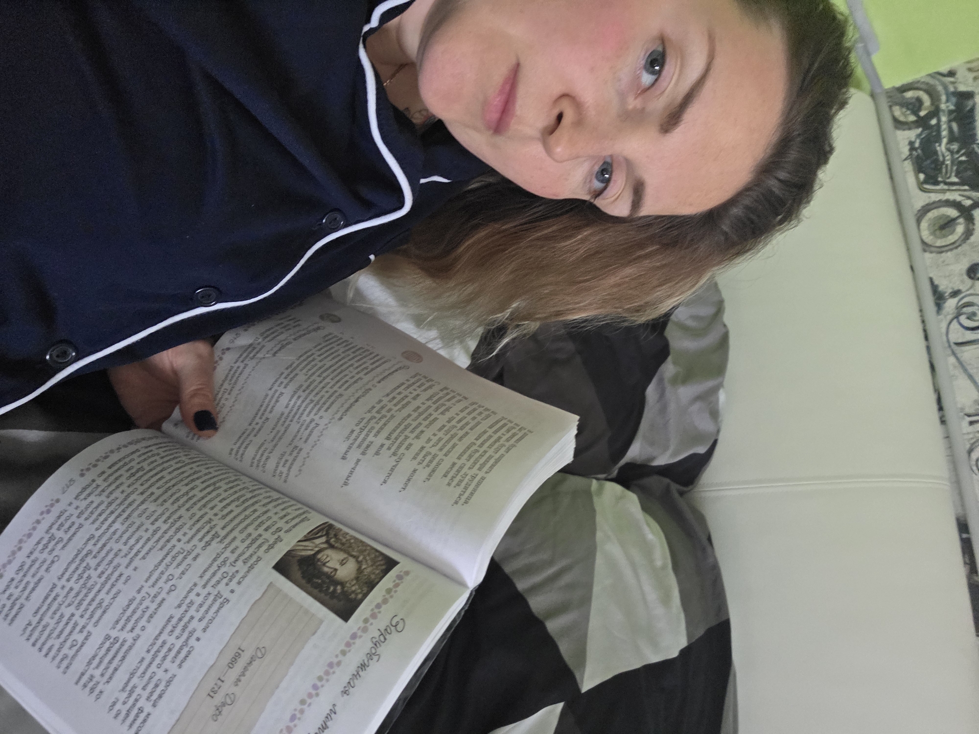 A woman is lying down on her side, reading an open book in an indoor setting, likely a bedroom or living room, in Chaikovsky, Russia. She is looking directly at the camera with a calm and relaxed expression. She has blue eyes, light brown hair, and fair skin, and is wearing dark blue pajamas with white piping. Her left hand, adorned with dark nail polish, holds the open book.
The book displays Cyrillic text on its pages. Clearly visible on the right page is a section about "Даниель Дефо" (Daniel Defoe), along with his birth and death years "1660-1731". Below this, there's a small, stylized illustration of a person. The text discusses Defoe's life, his background as a merchant's son, his disinclination to become a priest, his aspirations for travel, his endeavors in commerce and journalism, and the success of his first novel. A heading "Зарубежная лит" (Foreign Literature) is also visible.
The woman is resting on bedding with a grey and black striped pattern, next to a white upholstered surface that could be a headboard or couch cushion. In the background, a light green wall is visible, along with a patterned element on the right that resembles motorcycle or bicycle wheels.