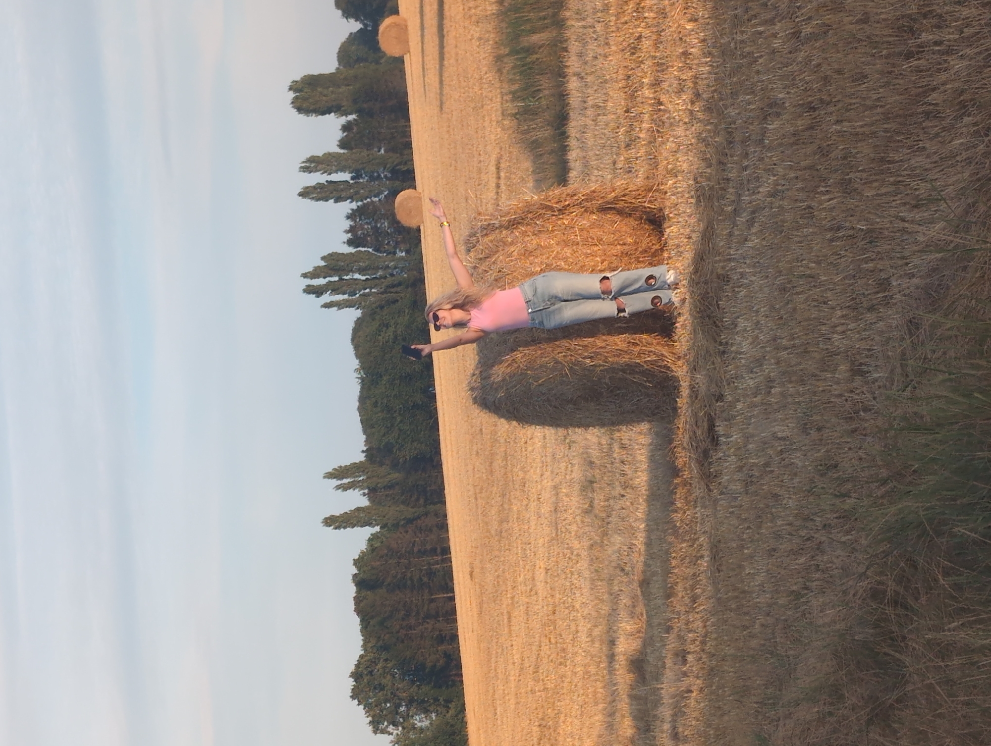 In Alvimare, France, a young woman with blonde hair stands in a field of dry, golden stubble under a pale blue sky with scattered white clouds. She is leaning against a large, round bale of hay, her left arm extended upwards, holding a black smartphone as if taking a selfie. Her right arm is also raised, open to the side. She wears a bright pink sleeveless top and distressed light blue jeans with large cutouts on the knees. The field is dotted with several other hay bales, and a line of trees forms the background, with some taller, slender trees interspersed with denser foliage. The lighting suggests it is late afternoon or early evening, casting long shadows and illuminating the scene with a warm, golden hue.