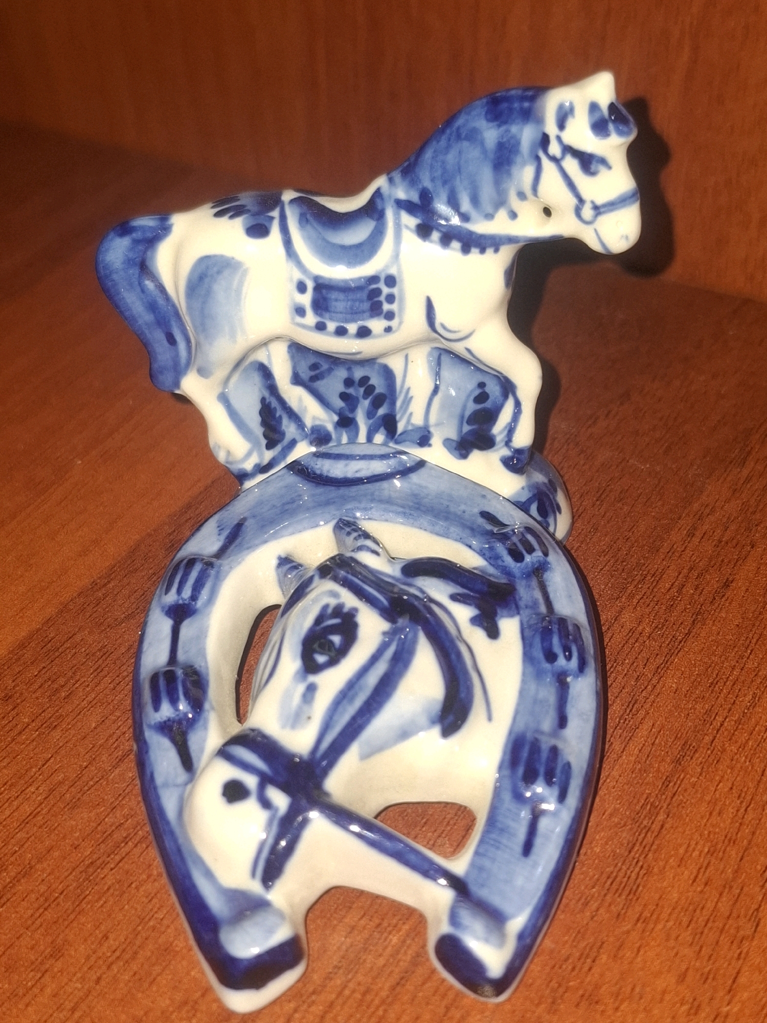 A close-up, overhead shot shows a ceramic decorative piece featuring two horses and a horseshoe. The objects are white with blue patterns, possibly indicative of Gzhel pottery. The lower portion of the piece shows a horseshoe with a horse's head emerging from it, depicted with blue eyes and bridle. Above this, a second horse stands on a raised platform, also adorned with blue patterns, including a saddle and intricate detailing on its body and legs. The background is a warm, reddish-brown wooden surface, likely a table or shelf, with subtle variations in grain and texture. The lighting casts a soft shadow behind the ceramic figures, suggesting an indoor environment. The image does not depict any location in Almaty, Kazakhstan.