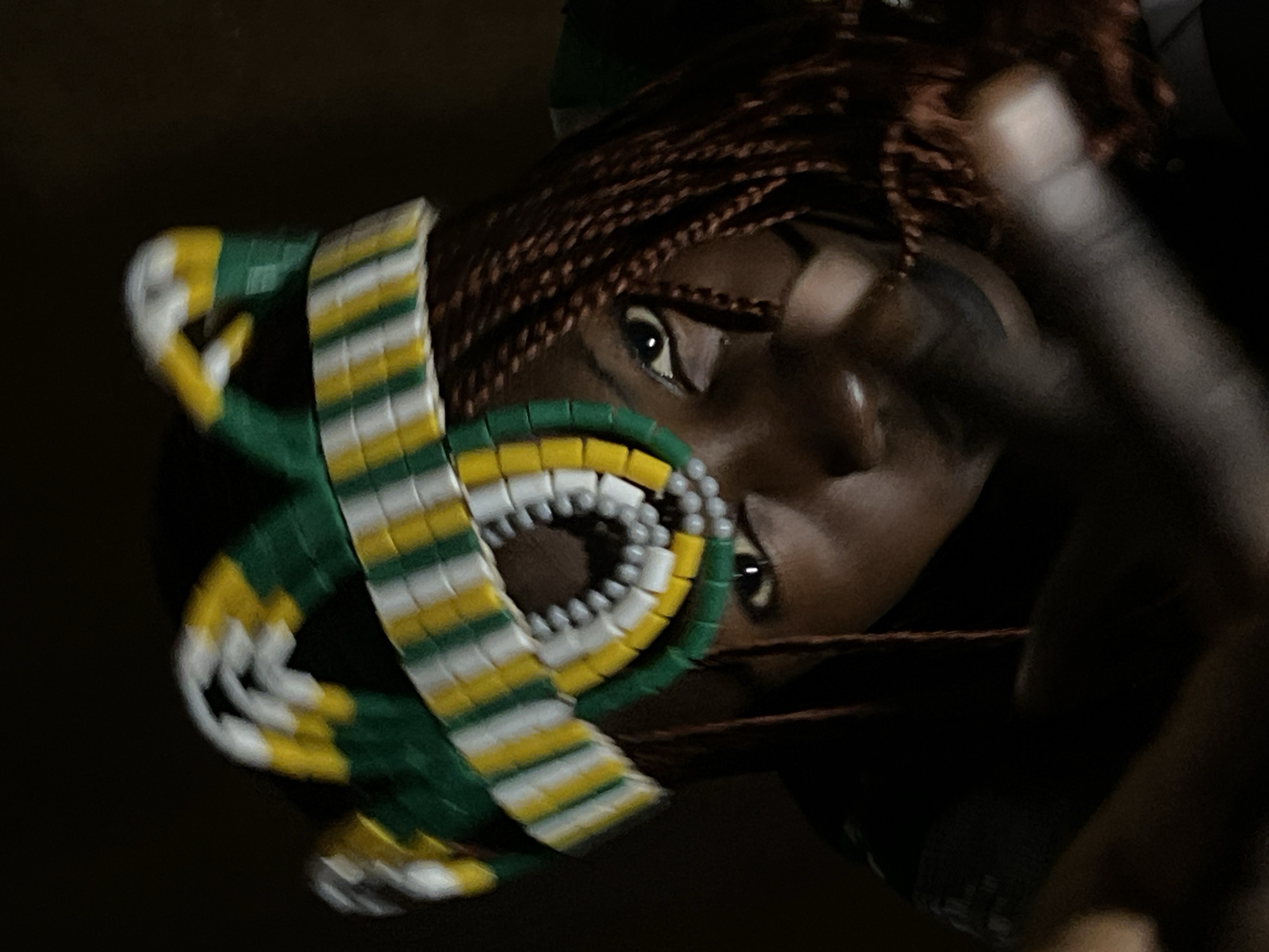 In a low-light, indoor setting, a young Black individual wearing a beaded headdress with yellow, green, and white stripes is the central subject. The headdress is elaborate, featuring a layered, crown-like design with beaded accents around the forehead. The individual's hair is braided into dark brown strands, some of which fall forward. The person's face is partially obscured by shadow, but their dark eyes are visible, looking towards the camera. Their lips are dark, possibly accentuated with makeup. The background is dark and indistinct, suggesting an unlit or dimly lit room. The image is a close-up, focusing on the individual's upper body and the headdress. There are no discernible architectural features that would definitively place the scene in Jos, Nigeria; the provided crops do not offer any environmental clues to support this location.
