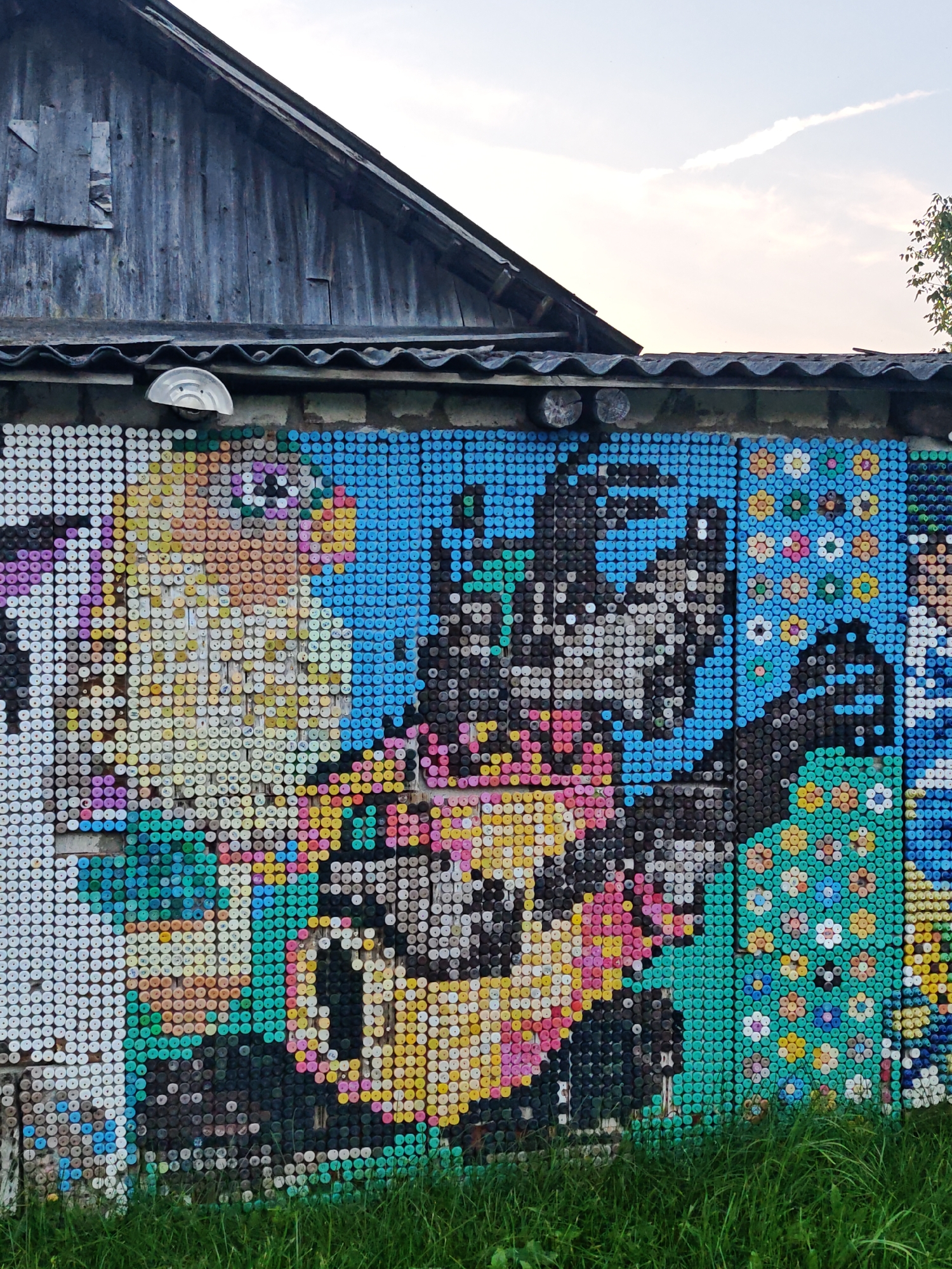 The image shows a section of a weathered wooden building with a sloped roof, situated behind a wall decorated with a mosaic made from plastic bottle caps. The mosaic depicts a colorful portrait of a person with distinctive features, including large eyes and rosy cheeks. The background of the mosaic features a blue sky and a pattern of small flowers in various colors against a turquoise backdrop. In the foreground, lush green grass grows at the base of the wall. The overall impression is that of an outdoor art installation, possibly in a rural setting.