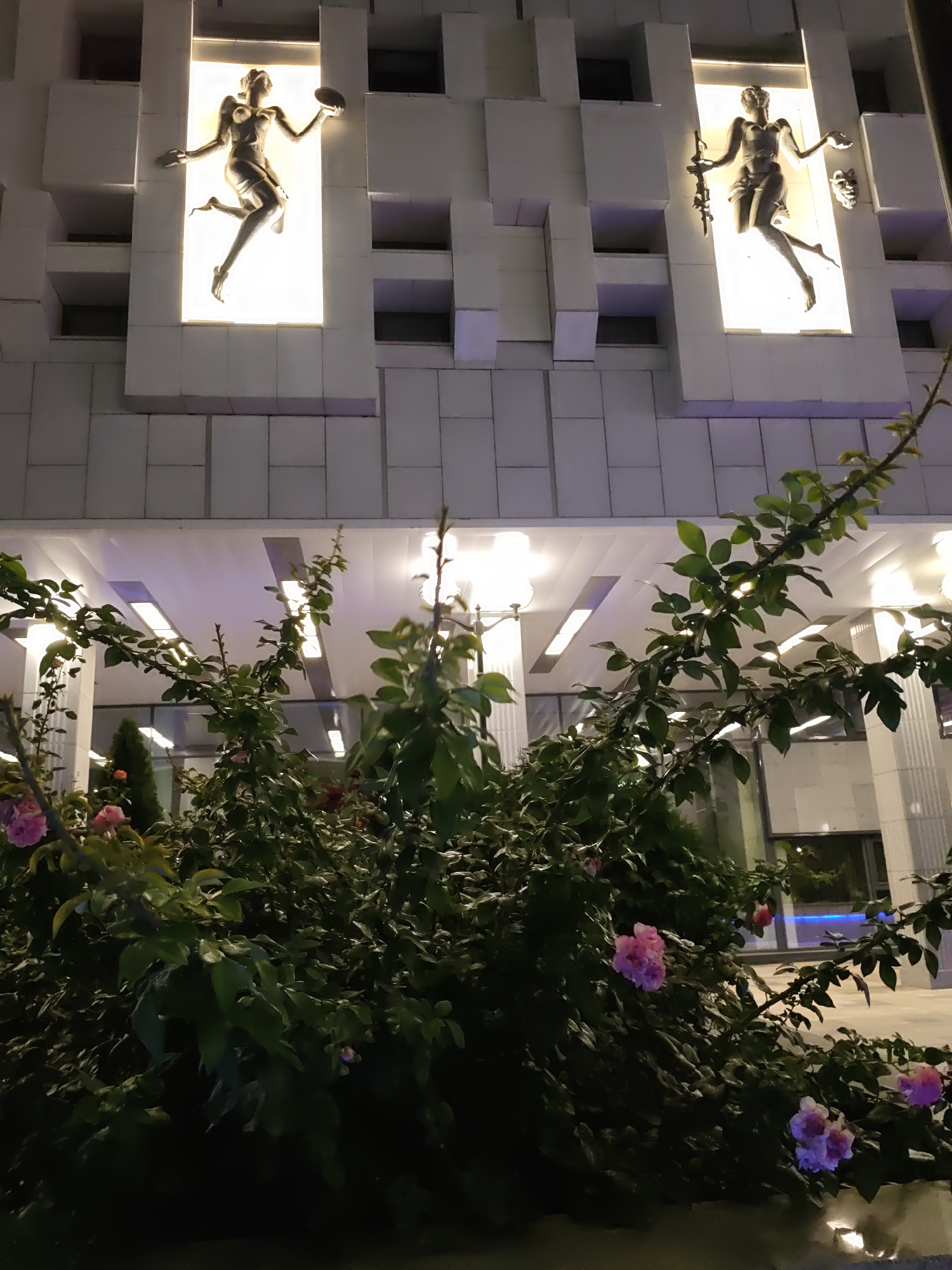 An exterior night shot shows the facade of a building adorned with two illuminated niches, each containing a metallic sculpture of a female figure. The figure on the left appears to be holding a disc in one hand and a smaller object in the other, while the figure on the right holds what looks like a staff. The building's exterior is constructed of white, rectilinear blocks arranged in a staggered pattern. In the foreground, a dense bush with dark green leaves and several pink flowers is visible, partially obscuring the lower portion of the building. The lighting is a mix of the bright white light emanating from the niches and the softer, purplish light from the building's interior or architectural lighting.