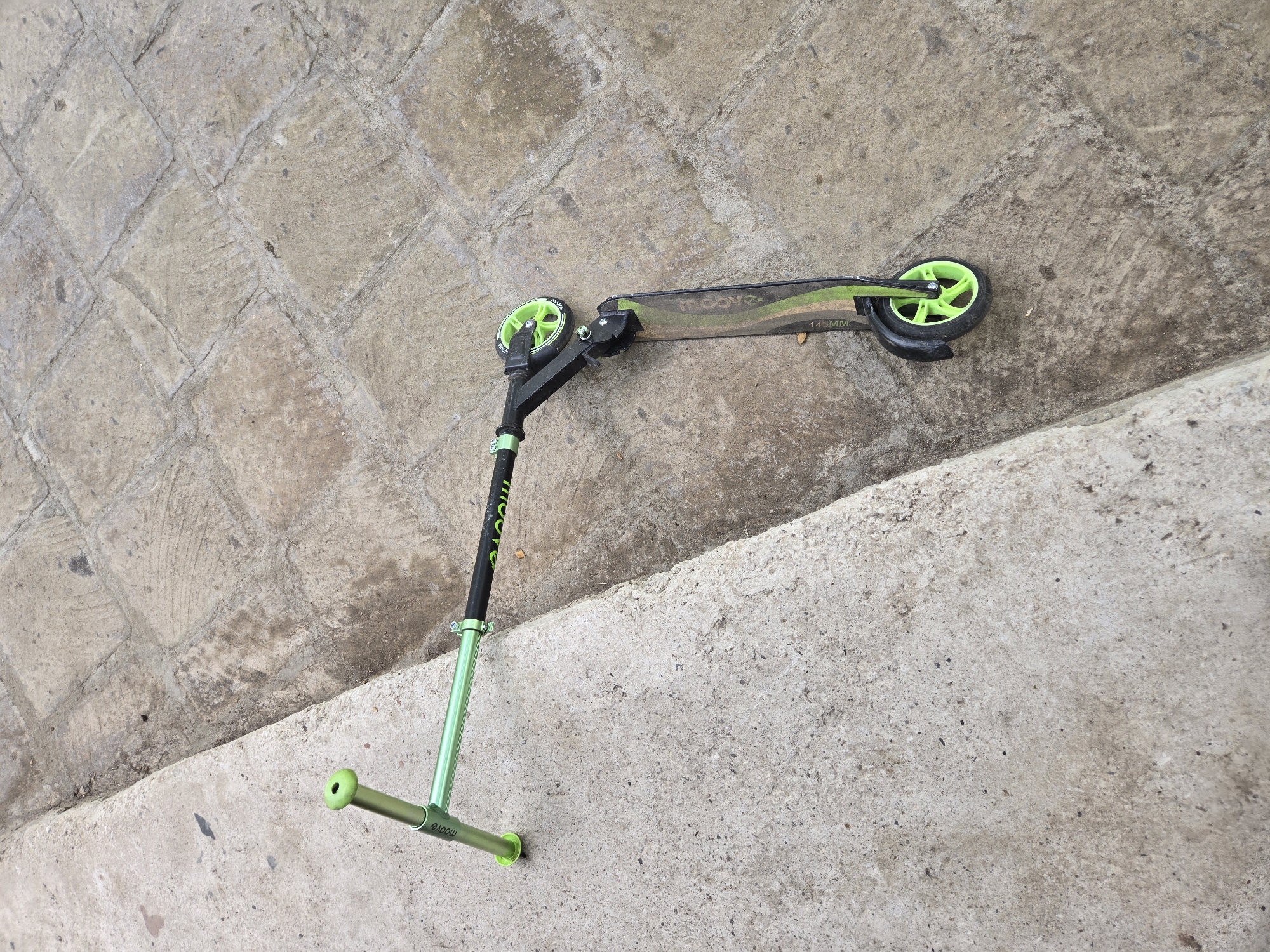 A green and black scooter lies on a concrete surface next to a raised concrete curb. The scooter is angled with its handlebars on the left and wheels on the right. The handlebars are a bright green with a black grip. The stem of the handlebars is also green and has the word "moove" printed on it. The scooter has two wheels, both with bright green spokes and black tires. The deck of the scooter is black with green stripes and text. The background consists of rough, textured concrete pavement.