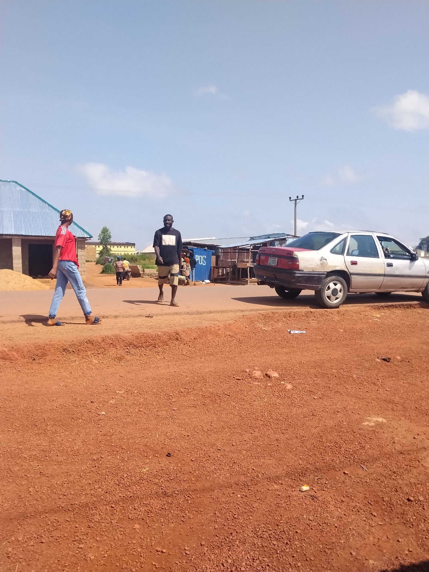 A wide shot captures an outdoor scene on a sunny day in Durumi, Nigeria. The foreground is dominated by reddish-brown dirt and gravel, indicative of unpaved ground. In the mid-ground, a paved road runs horizontally, separating the dirt from a collection of structures. On the left side of the road, a building with a corrugated metal roof is under construction, with piles of sand beside it. Further down the road, a white sedan with a red trunk is parked. Two men are walking on the paved road. One, wearing a black t-shirt and patterned shorts, is walking towards the camera. Another, in a red t-shirt and blue jeans, is walking away from the camera. In the background, several stalls with corrugated roofs are visible, and a blue container with "POS" written on it is present. A power pole with wires extends into the sky. Several other people and a motorcycle are visible in the distance, further down the road. The sky is blue with scattered white clouds.