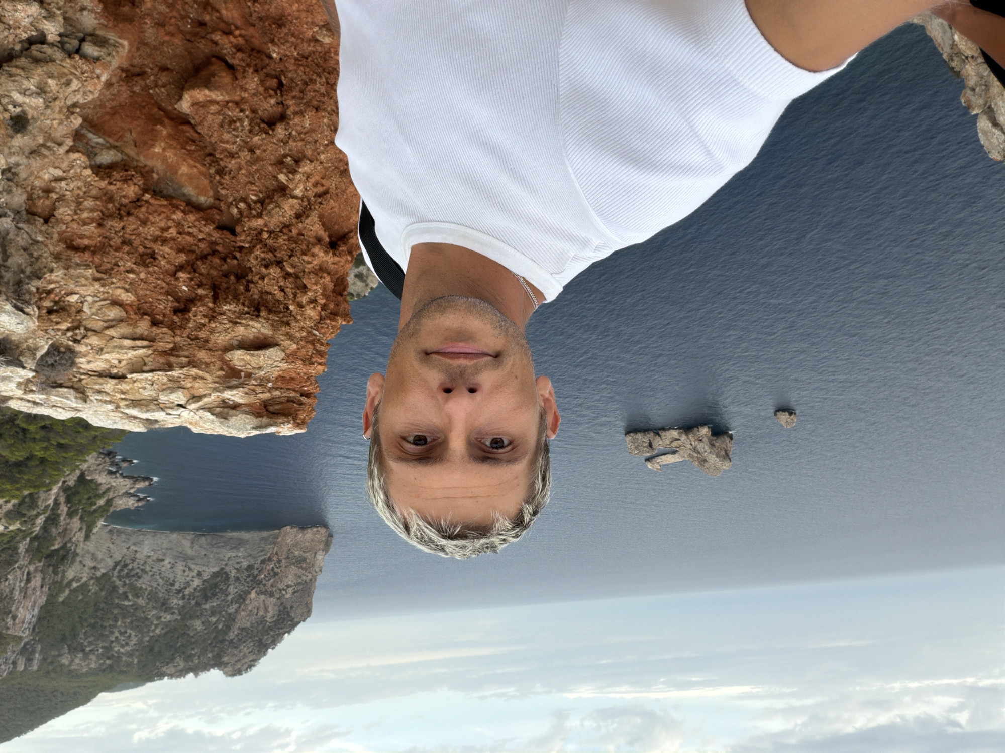 This is a selfie taken by a person with blonde hair in the city of Sant Antoni de Portmany, Spain. The person is wearing a white t-shirt. They are standing on a cliff overlooking the ocean. In the background, there are islands and cliffs. The sky is cloudy.