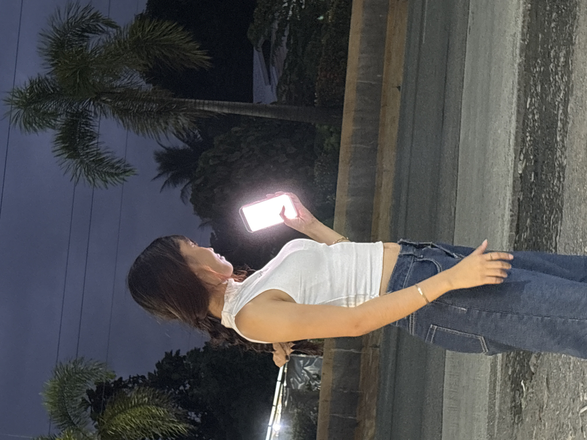 The image captures a young woman standing on a street at night, illuminated by the bright screen of her smartphone. She is dressed in a white sleeveless top and blue denim jeans. Her hair is dark and long, pulled back into a braid. The background features palm trees, vegetation, and a dimly lit street. The setting appears to be an outdoor urban environment, likely a street in Carmen, Philippines, given the presence of tropical vegetation and the general atmosphere. The lighting suggests it is evening or nighttime, with artificial lights from the phone and possibly distant sources.