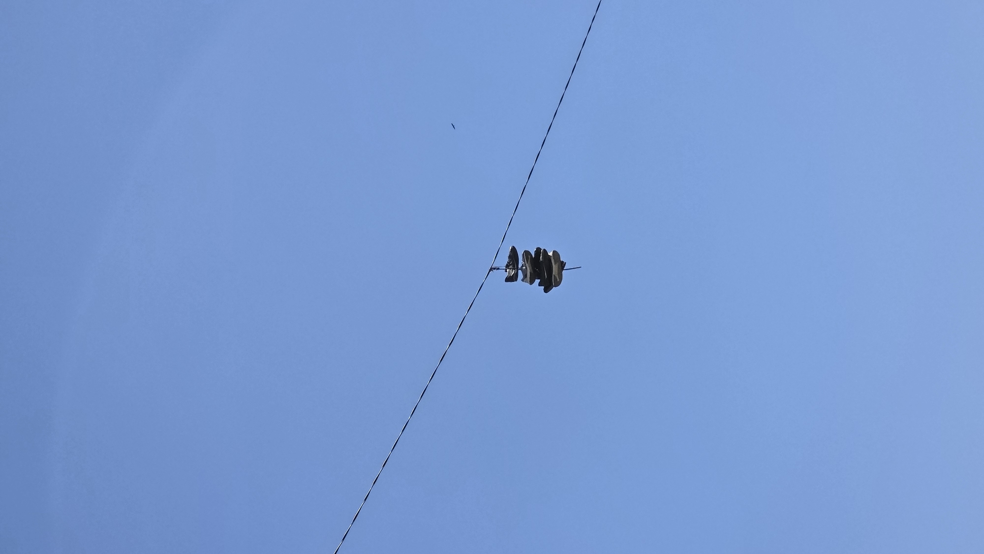 Against a clear blue sky, a pair of shoes are suspended from a power line. A bird is also visible in the sky. The shoes appear to be dark in color and are hanging by their laces. The power line runs diagonally across the frame. The scene is consistent with urban areas where shoes are sometimes thrown onto power lines. Specific location is unknown.