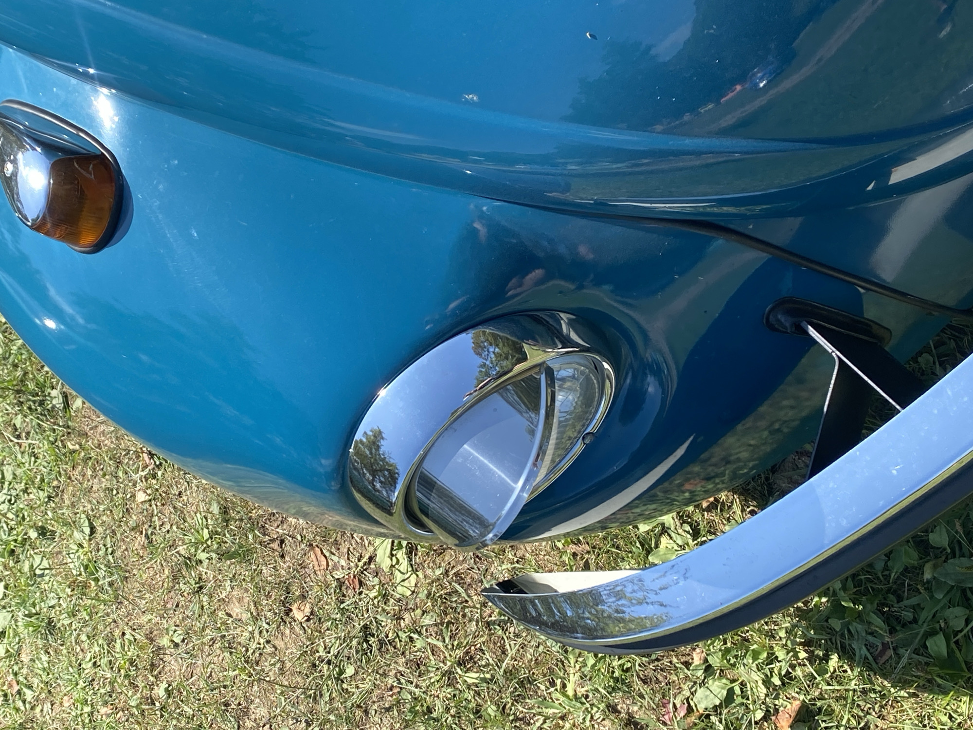 The image shows the front quarter of a vintage turquoise Volkswagen Beetle parked on a grassy area, likely in Bezymyannoe, Russia. The car's distinctive rounded headlight with a chrome bezel is prominently featured. A chrome bumper extends across the lower part of the image. A small amber turn signal is visible on the hood. The grass is dry and appears to be patchy in places, suggesting a warm, sunny environment. No people or other vehicles are visible in the frame.