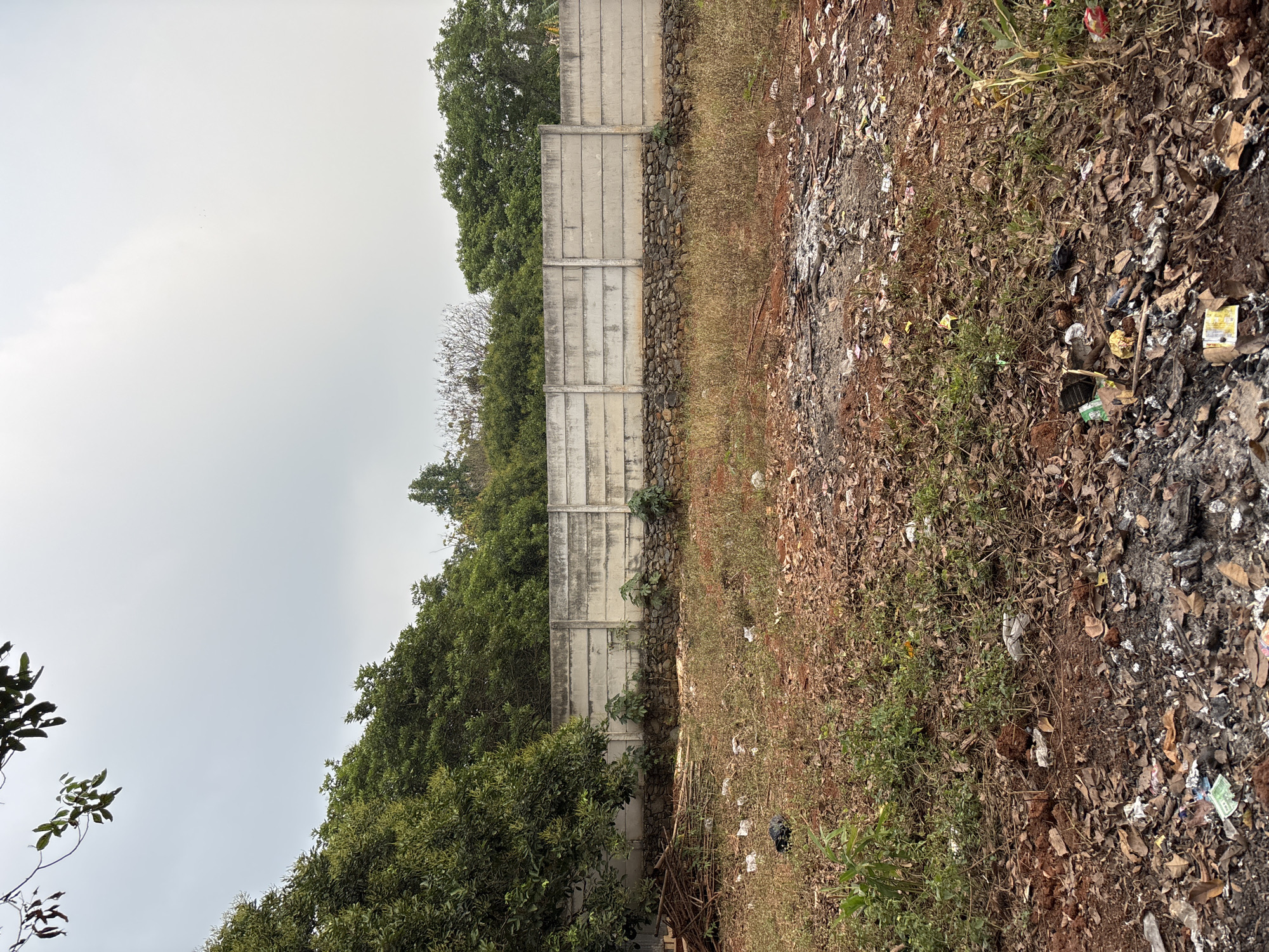 In Taman Rahayu, Indonesia, a concrete barrier wall stands prominently, separating a debris-strewn lot from a dense green foliage backdrop. The wall, composed of precast concrete panels, is erected on a foundation of rough-hewn stones. Overgrown vegetation, including dry grasses and scattered leaves, covers the ground in the foreground, interspersed with discarded plastic bags, wrappers, and other litter, suggesting a state of neglect. A pile of rusted metal and wooden debris is visible to the left of the wall, further contributing to the unkempt appearance of the scene. The sky above is overcast, casting a diffused light on the landscape.