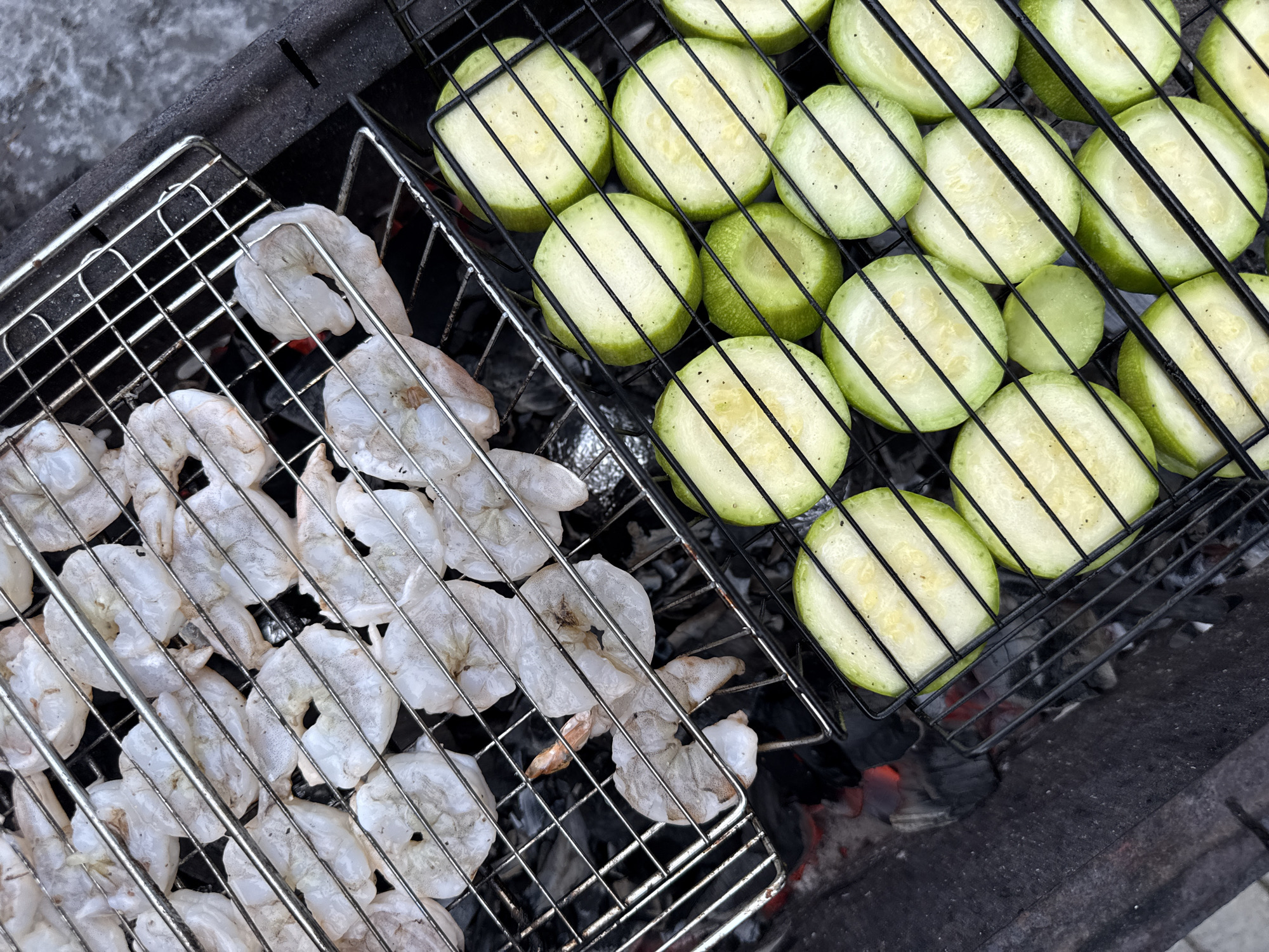 The image shows two grill baskets on top of a barbecue grill. One basket contains peeled shrimp, and the other contains sliced zucchini. The grill is situated outdoors and based on known information, the location can be presumed to be Votkinsk, Russia. The shrimp appear to be raw, and the zucchini slices are arranged in an orderly fashion. Coals are burning beneath the grill baskets.
