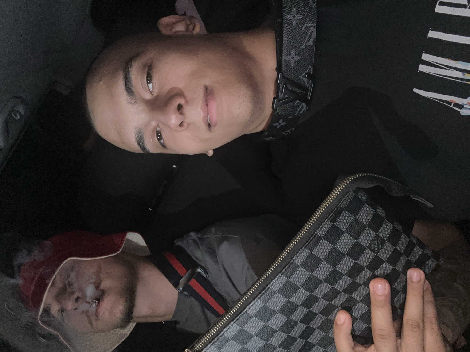 The image depicts two men in a dark environment, likely a car interior. One man is positioned at the top, facing the camera with a neutral expression. He is wearing a black shirt with white designs and a black Louis Vuitton strap across his chest. The other man is below him, partially obscured by smoke emanating from his mouth, presumably from a cigarette or joint. He is wearing a bucket hat with red and white. A Louis Vuitton Damier Graphite pattern clutch or bag is held by a hand in the foreground. No location in the city of Plovdiv, Bulgaria is evident.