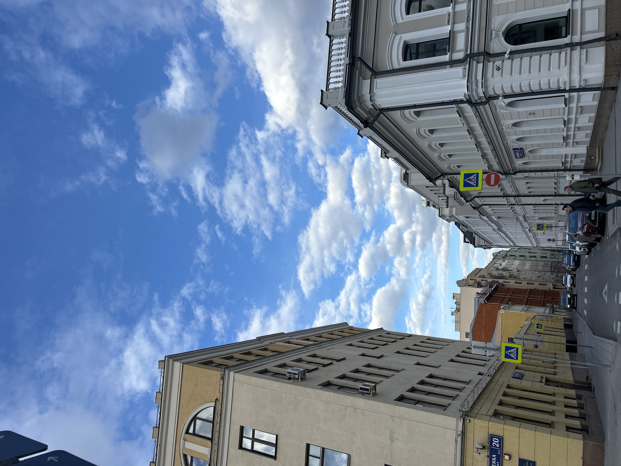 The image depicts an urban street scene in an unidentified city. Buildings line both sides of the street, with varying architectural styles and heights. The sky is predominantly blue with scattered white clouds. On the right side of the street, pedestrians are visible. Cars are parked along the street. A yellow pedestrian crossing sign is mounted on a pole near a building on the left side of the street. A building address sign is located at the lower right.