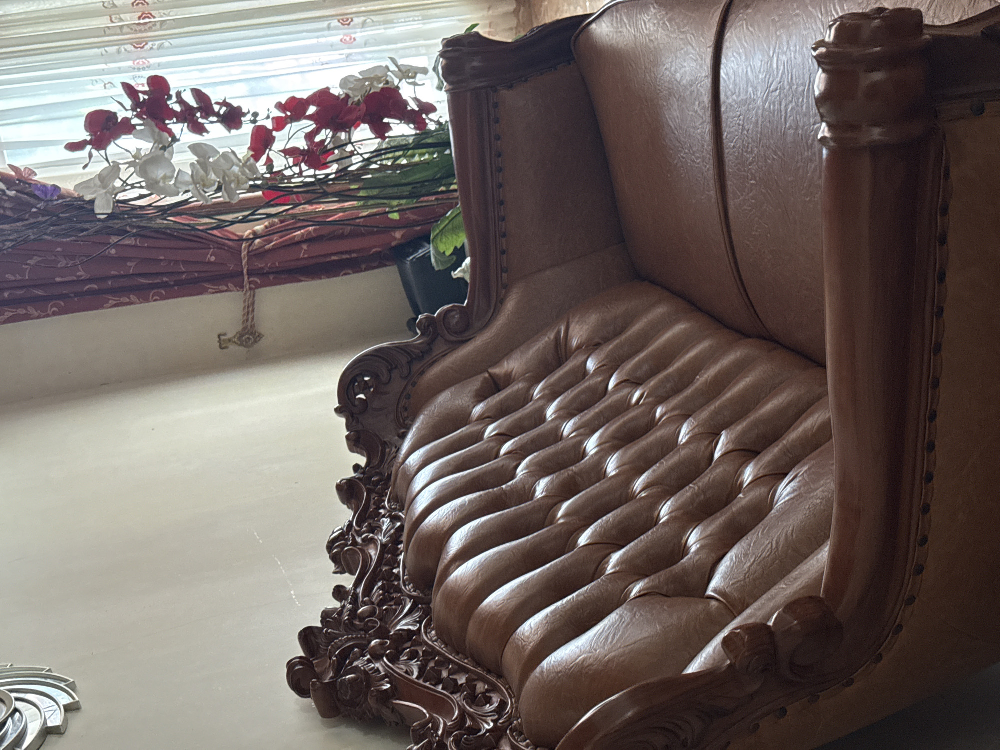A brown leather tufted sofa with ornate dark wood trim is the main subject of the image, captured in a room. The sofa has a deeply tufted backrest and arms. To the right of the sofa, a window is visible with sheer white curtains and a darker red curtain tied back. In front of the window, a decorative arrangement of artificial red and white orchids on branches is displayed. The room appears to be a living area, with a plain wall and a partially visible wall decoration on the left. The lighting suggests an indoor setting with natural light coming from the window. The scene is static and appears to be a still life composition. No individuals are present in the image. The specific location in Bireuenmeunasahblang, Indonesia is not visually ascertainable from the image itself.