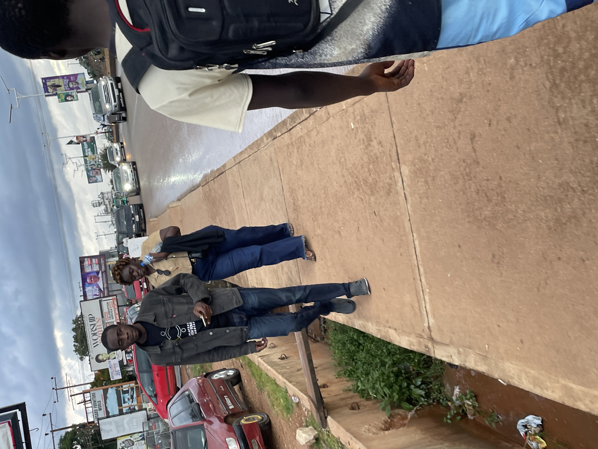 In Jos, Nigeria, an overcast sky looms over a street scene. A man in a dark jacket and jeans walks on a reddish-brown concrete sidewalk, holding a mobile phone. Behind him, a woman in a tan shirt and blue jeans walks, carrying a dark bag and a water bottle. A red car is parked on the left side of the frame, and another red car is further down the street. The sidewalk borders a shallow drainage ditch filled with green vegetation and some debris. In the background, billboards line the opposite side of the street, and several vehicles, including a silver SUV, are driving on the wet pavement. Another person, partially visible, walks towards the camera on the right side of the sidewalk, wearing a light-colored t-shirt and blue shorts, with a backpack on.