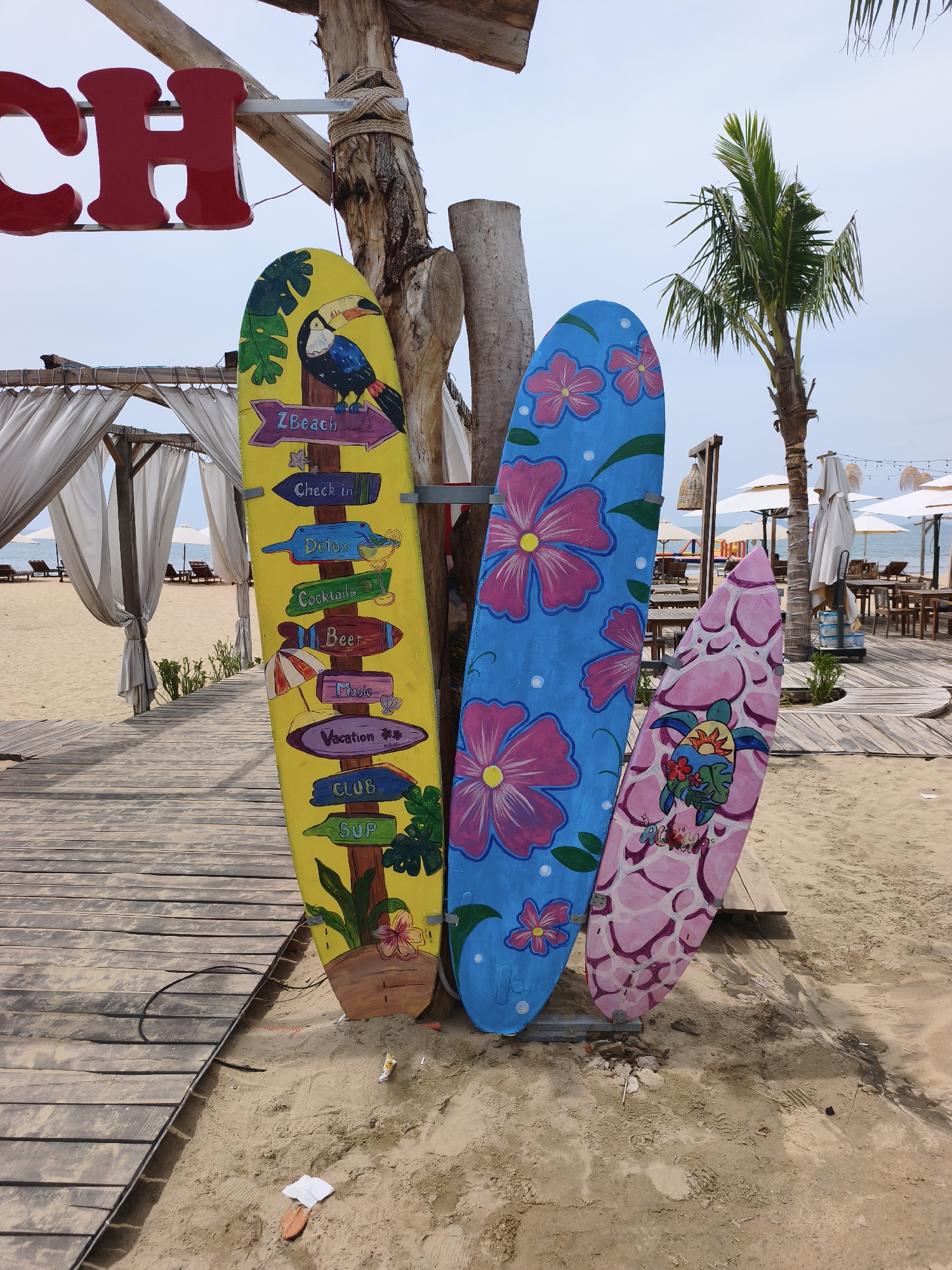 Three decorative surfboards stand upright in the sand near a wooden boardwalk at a beachside establishment in Quy Nhon, Vietnam. The surfboard on the left is yellow with painted signs pointing to various amenities like "ZBeach," "Check in," "Detox," "Cocktails," "Beer," "Music," "Vacation," "Club," and "SUP." It also features a painted toucan and tropical leaves. The middle surfboard is bright blue with large pink floral designs. The surfboard on the right is pink with a lighter pink pattern and features a painted sea turtle wearing a flower lei. Behind the surfboards, a wooden structure with white canopy tents and lounge chairs is visible, along with a palm tree and tables and chairs for an outdoor dining area. The beach and the ocean are in the background. The sky is overcast.