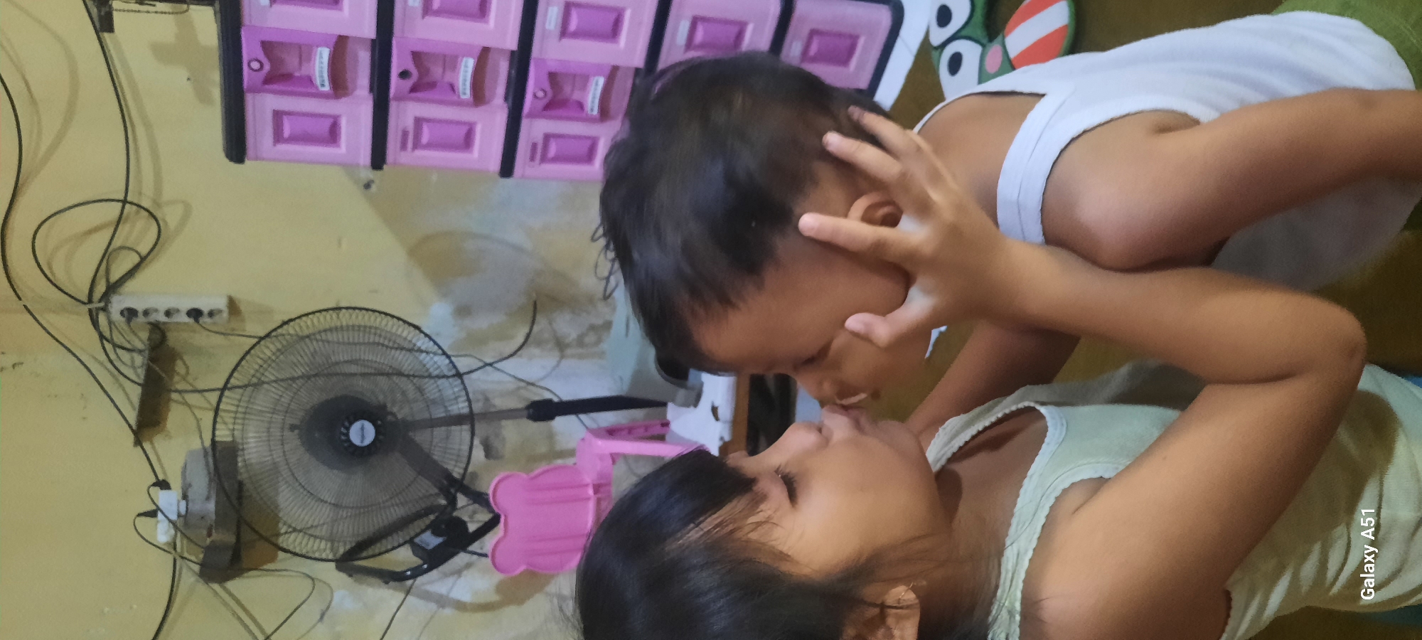 Two young children are shown in a close-up shot, their faces almost touching as if about to kiss. The child on the left, with dark hair and closed eyes, wears a light green tank top. The child on the right, also with dark hair, is wearing a white tank top and has their hands raised, gently touching the other child's face. The background is dimly lit, revealing a fan, a pink child-sized chair, and a pink plastic storage cabinet. The environment appears to be indoors, possibly a residential setting in Ploso, Indonesia, given the presence of various household items and the overall rustic ambiance. The image is a candid moment capturing an intimate interaction between the two children.