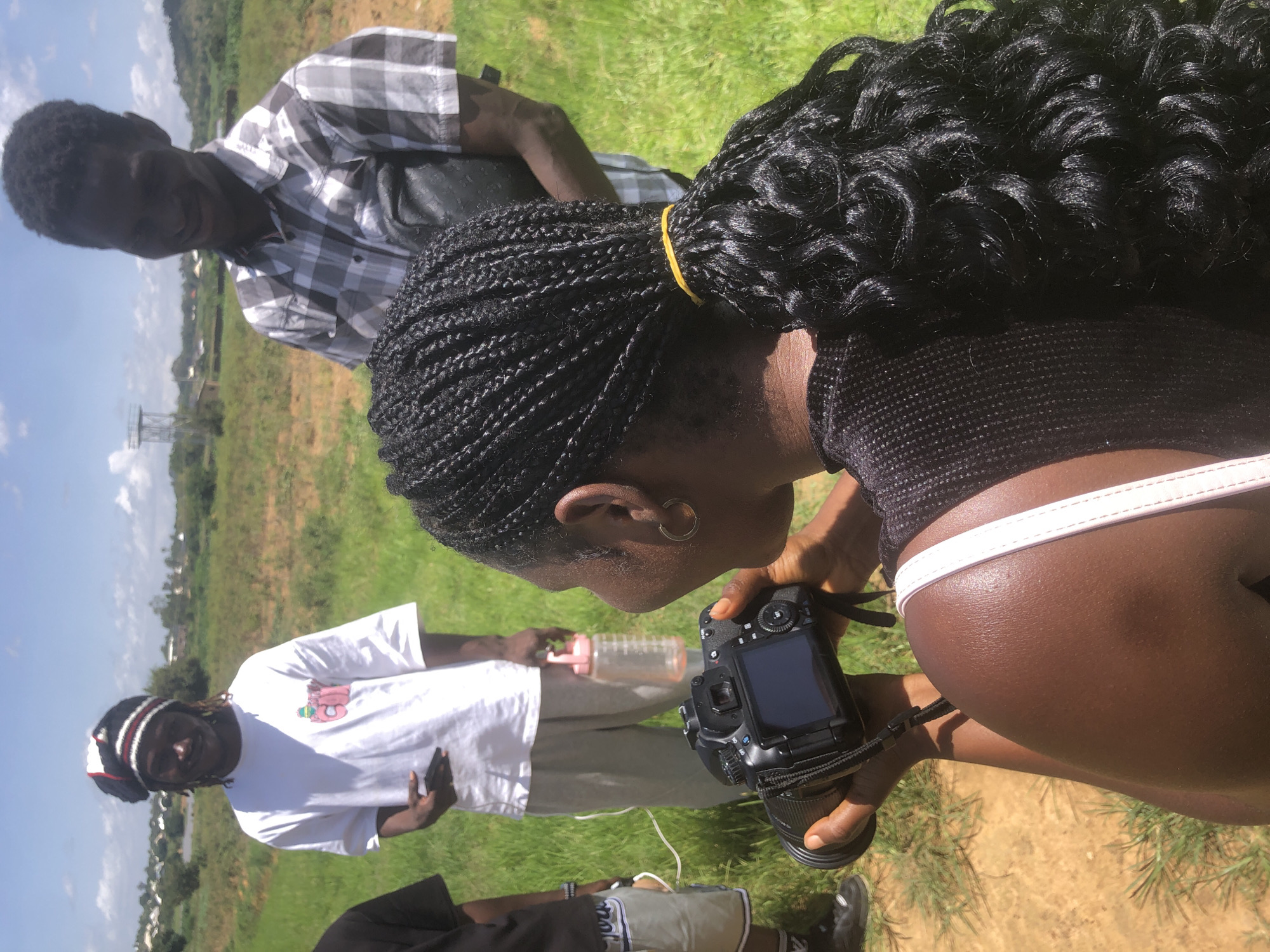 In a grassy field under a bright sky, a group of four individuals is gathered. The primary focus is a woman from the shoulders up, holding a black DSLR camera with her right hand on the lens and her left hand supporting the camera body. She has dark skin and her hair is styled in numerous tight braids, gathered into a ponytail at the back of her head with a yellow elastic band. She is wearing a black top with a mesh-like texture.
To her left, a man is visible from the waist up. He has dark skin and is wearing a checkered short-sleeved shirt in shades of grey and white. He is holding a dark bag over his left shoulder and appears to be looking down and smiling.
Behind them, another man is standing, seen from the torso up. He is wearing a white t-shirt with a colorful graphic and a striped knit beanie. He holds a black smartphone in his left hand. Beside him, and slightly in front, a pink plastic water bottle with a white cap is visible.
A fourth person is partially visible on the far left, only showing their legs and lower torso. They are wearing light-colored shorts with a blue stripe and dark sneakers.
The environment is an open, grassy area with some patches of bare earth. In the background, there are trees and buildings suggesting a semi-rural or suburban setting, possibly on the outskirts of Gwafan, Nigeria. The ground in the foreground is a mixture of reddish-brown earth and green grass. The sky is blue with scattered white clouds.