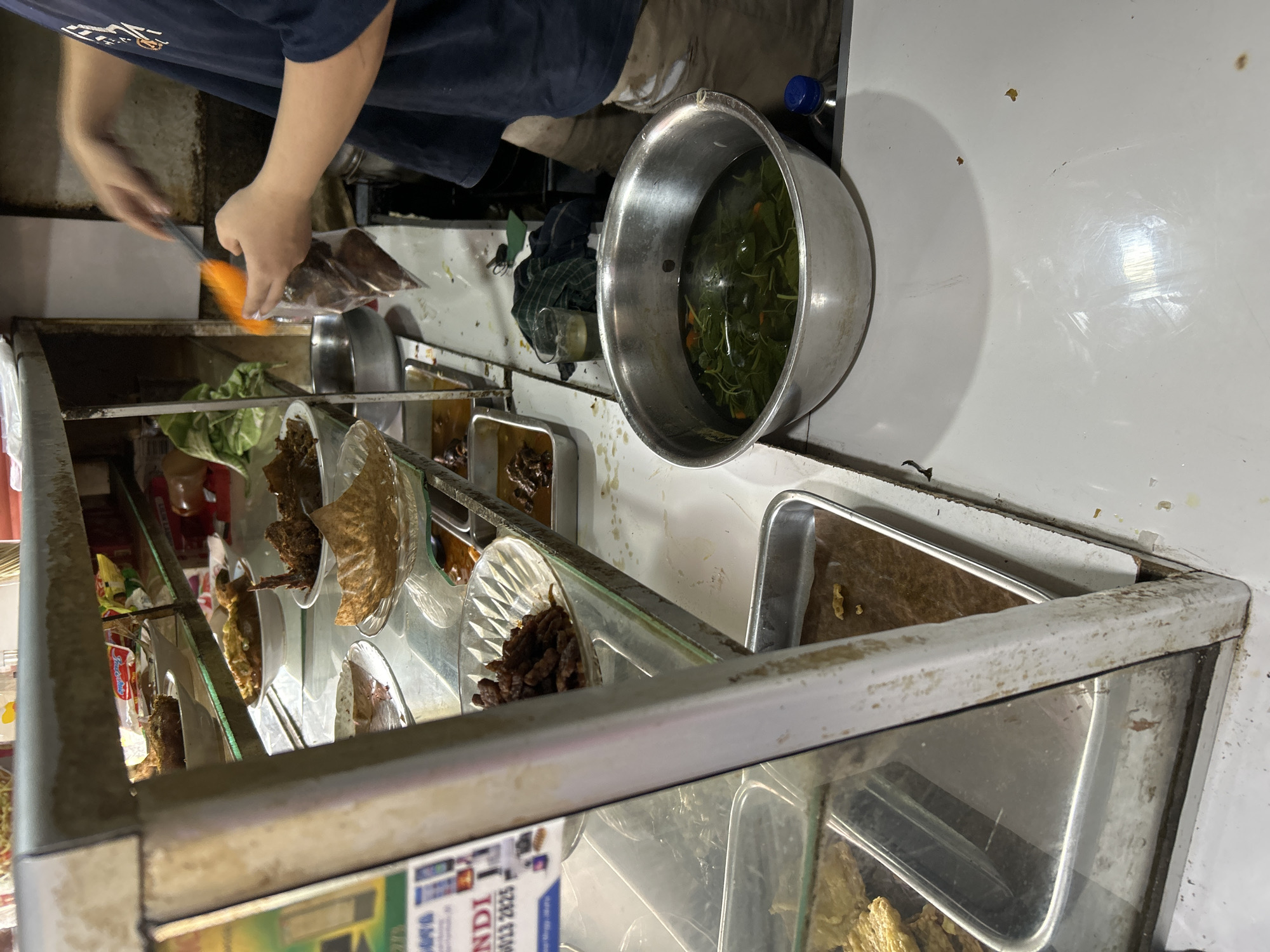 A person is preparing food in a market stall in Depok, Indonesia. The stall appears to be open-air or semi-enclosed, with a metal shelving unit displaying various food items in containers. The person, wearing a dark blue t-shirt and light-colored apron or shorts, is seen with their hands holding a plastic bag and a utensil, seemingly portioning out food. In the foreground, a large metal bowl contains a green leafy vegetable soup with pieces of orange. Behind the bowl, a clear glass contains a light-colored liquid, and a dark-colored towel is draped nearby. The shelves above display an assortment of dishes, some appearing to be fried items, curries, and rice. Trays of food are also visible in a refrigerated display case to the left. The overall environment suggests a busy food vendor setting, typical of Indonesian street food markets.