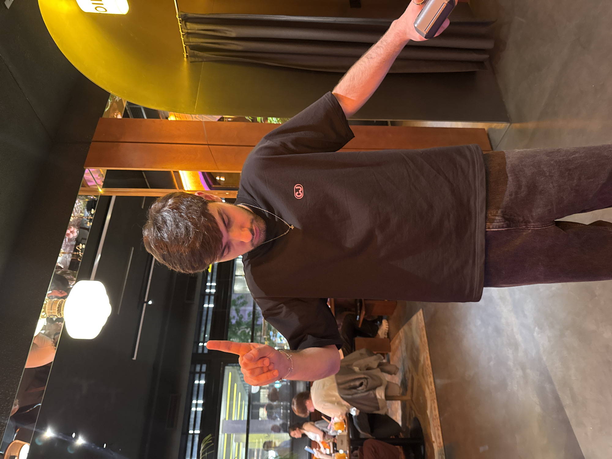The image captures a man indoors, seemingly at a restaurant. The location appears to be in Yerevan, Armenia.
The man, who has brown hair and a beard, is wearing a black t-shirt with a small, circular logo on the left side of his chest. He is pointing forward with his right hand, which has a bracelet on the wrist. He also has a thin necklace on. His left arm is raised, holding an object that is only partially visible. He is looking down in the direction that he is pointing. He is wearing dark-colored pants.
The setting includes restaurant elements, with tables, chairs, and other patrons visible in the background. There's a reflective surface to the left, perhaps a mirror or glass, showing a glimpse of the restaurant's lighting and decor. The lighting appears to be a mix of natural light from outside and artificial light from the fixtures within the restaurant.
The foreground shows the floor. The background features the restaurant interior, windows with outside views, and other seated individuals who are engaged in various activities, such as drinking and conversation.
