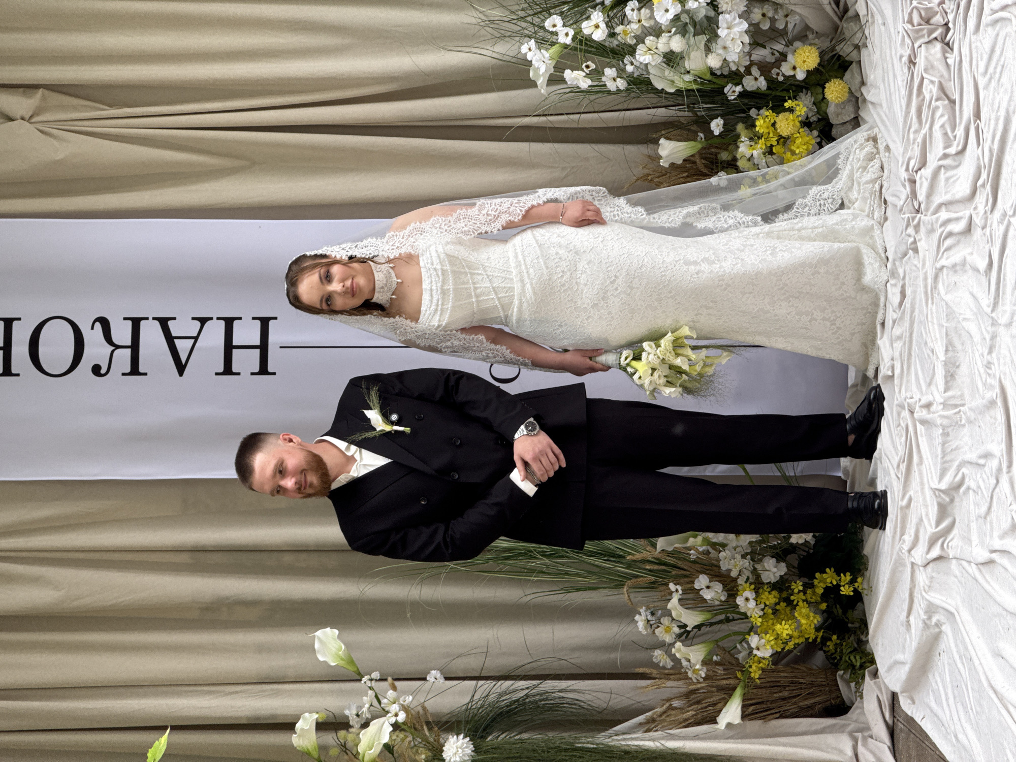 This full-shot vertical image captures two individuals, a man and a woman, standing side-by-side in a staged setting. The specific location within Perm, Russia, is not identifiable from the visual evidence.
The man, positioned on the left, is in his late 20s to early 30s with a short haircut and a trimmed beard. He wears a black double-breasted suit jacket, an open-collared white shirt, black trousers, and black dress shoes. A white calla lily boutonnière is pinned to his left lapel, and a silver-toned watch is on his left wrist. His hands are clasped in front of him as he looks towards his right (viewer's left) with a slight smile.
The woman, on the right, appears to be in her early to mid-20s. She is dressed in a white, floor-length lace wedding gown featuring a strapless sweetheart neckline and a corset-style bodice. A long, sheer white veil with lace trim extends down her back. She wears a white beaded choker necklace, a bracelet on her left wrist, and dark earrings. In her left hand, she holds a bouquet composed of white calla lilies, yellow flowers, and greenery. She maintains a neutral expression, looking directly forward.
The background consists of light beige or off-white draped fabric curtains, with a section on the upper right gathered. A tall, narrow white banner with black vertical text, partially visible as "НАКОР," stands between the individuals and the curtains. Elaborate floral arrangements featuring white calla lilies, various white and yellow flowers, and dried grasses are placed on both sides of the subjects. The floor is covered with a crumpled, light-colored fabric. The scene is evenly lit.