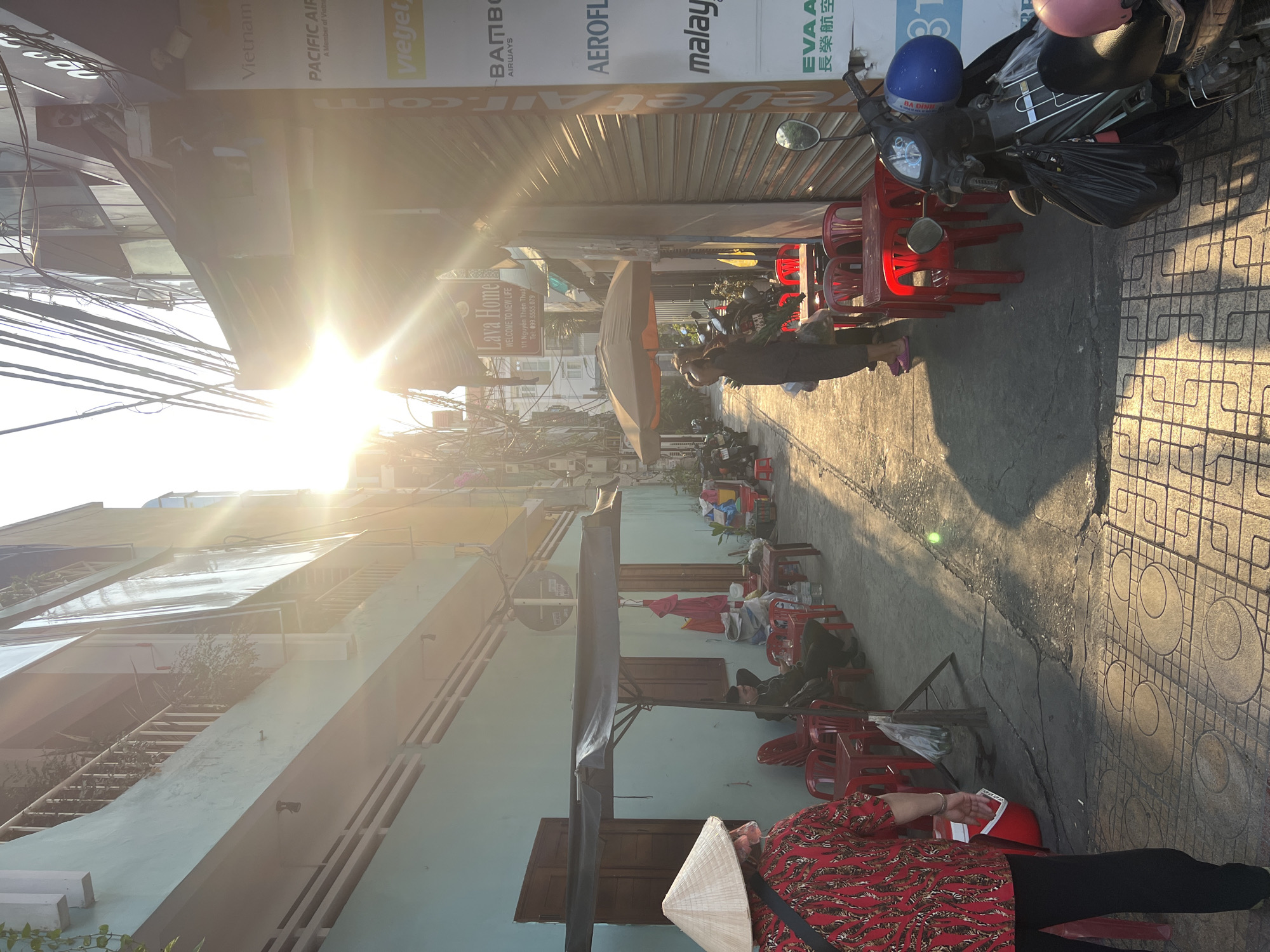 This outdoor scene depicts a street in Nha Trang, Vietnam during early morning sunlight hours. The street is lined with buildings on either side, identifiable by signage of airlines such as Pacific Air, Vietjet, Bamboo Airways, Aeroflot, Malaysia, and EVA Air. An establishment named Lava Home is also marked with a sign. People are seen along the street, some wearing traditional Vietnamese conical hats, red and black clothing, and black clothing. Red plastic chairs are placed along the street. Parked motorbikes are present and a tan umbrella is erected along the side of the street. Overhead cables cross the sky. The environment displays strong sunlight and shadows.