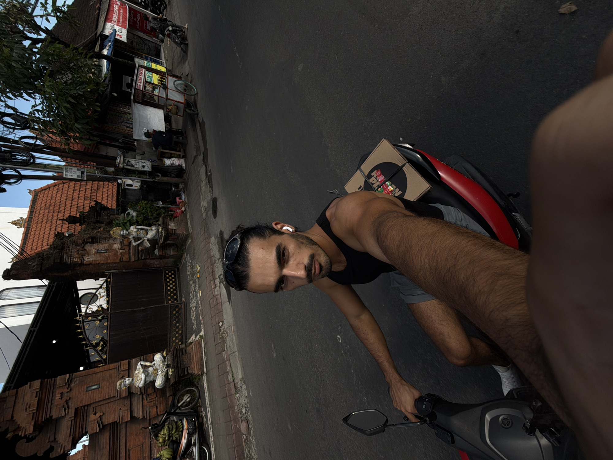 A man is captured in a selfie-style photograph while riding a scooter on a sunny day in North Kuta, Indonesia. The image is taken from a first-person perspective, with the man's right arm extended to hold the camera.
The primary subject is a man with dark hair tied back, wearing sunglasses pushed up on his head, a black tank top, and grey shorts. He has a beard and is looking directly at the camera with a neutral expression, wearing a white earbud in his left ear. His left hand is on the handlebars of a grey and red Yamaha scooter.
On the back of the scooter, a cardboard box is secured, likely for a delivery. The box features a black circular logo with white text, partially visible as "HAPPY FOOD", surrounded by smaller colorful squares or rectangles.
The background reveals a bustling street scene. On the left side of the road, there are traditional Balinese buildings with intricate wooden carvings and red-tiled roofs, adorned with white stone statues, possibly depicting deities or traditional figures. A dark metal gate is also visible. Further down the street, a small food stall or cart is present, with signs that clearly read "MIE AYAM BAKSO" (chicken noodle soup and meatballs), and partially visible text like "GADO SA..." and "ES TEH". Another red sign in the distance says "WARUNG" (small shop). Overhead, numerous electrical wires and cables crisscross. Trees with green foliage line the street. The road is asphalt, and the bright sunlight suggests daytime with clear weather. The overall setting strongly reflects the local culture and architecture of North Kuta, Indonesia.