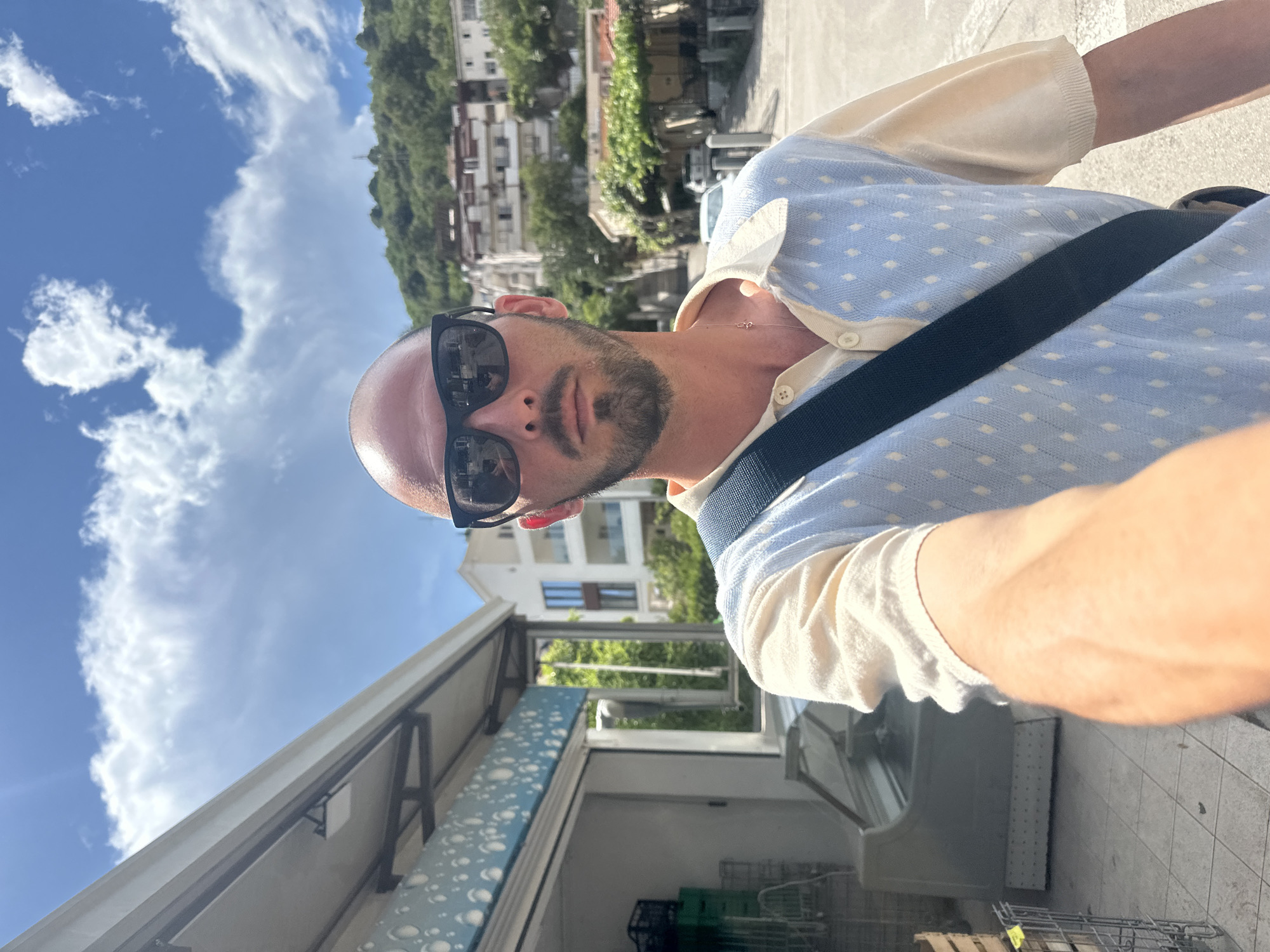 The image shows a person with a shaved head and a beard wearing sunglasses and a light-colored collared shirt with a blue patterned overlay. They are outdoors, with a clear blue sky and white clouds visible. In the background, there are buildings and lush green vegetation on a hillside, suggesting a Mediterranean or coastal setting. The person is carrying a dark bag across their chest. The perspective suggests a selfie taken from a slightly low angle. To the right, a part of a storefront or market stall is visible, with an open refrigerated display case inside. The overall impression is of a bright, sunny day in a European town. No text is visible in the image.