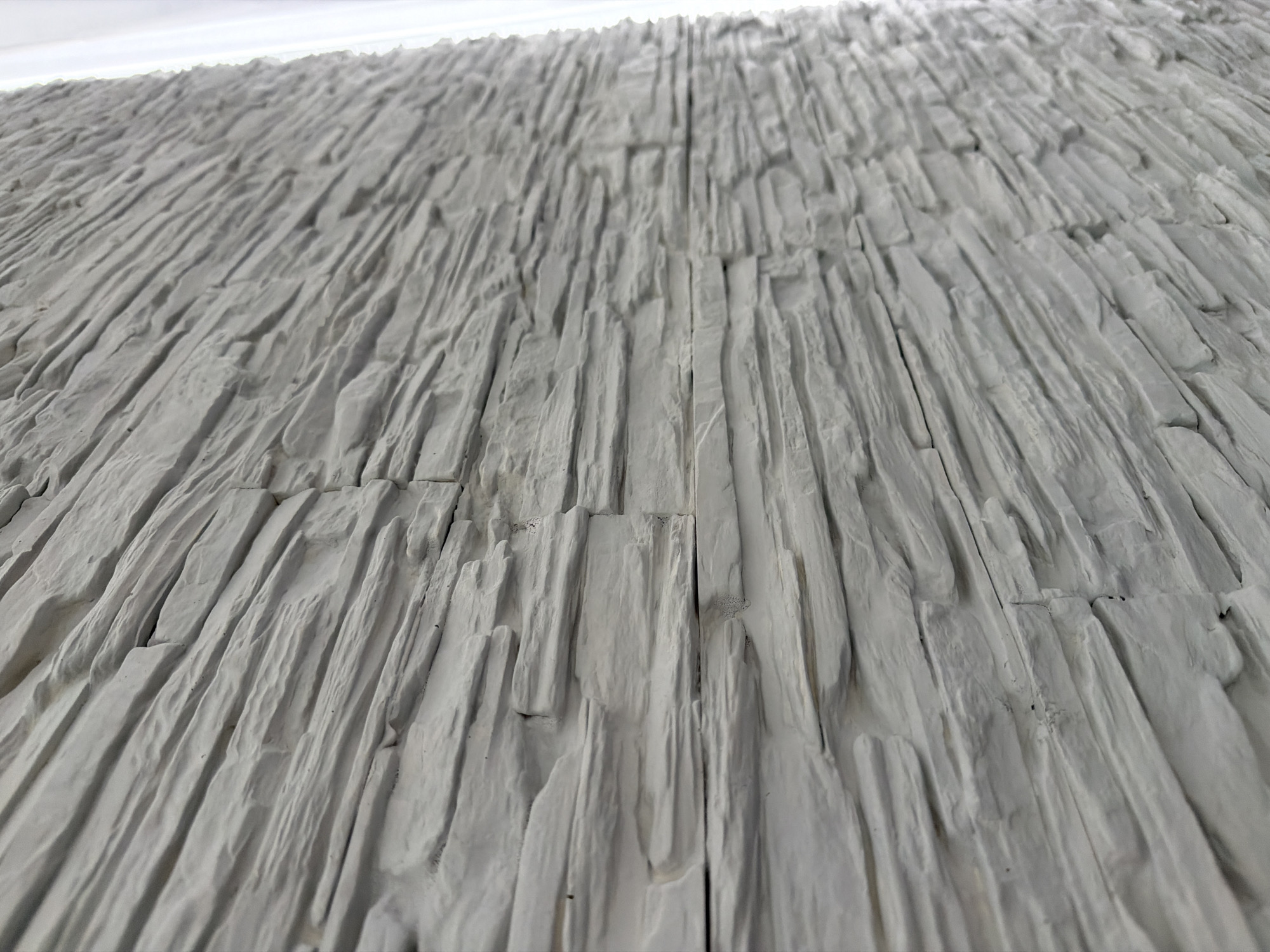 The image shows a close-up, right-facing, angled view of a wall covered in light gray, stacked stone veneer panels. The panels have a rough, irregular texture, mimicking natural slate or rock. The lighting appears to be from the right, casting subtle shadows that highlight the textured surface. There is a vertical element, possibly a window frame or curtain, visible on the far right edge of the image, which is brightly illuminated. The wall extends diagonally into the background, suggesting it is part of a larger architectural feature. The scene is indoors, indicated by the controlled lighting and the absence of outdoor elements.