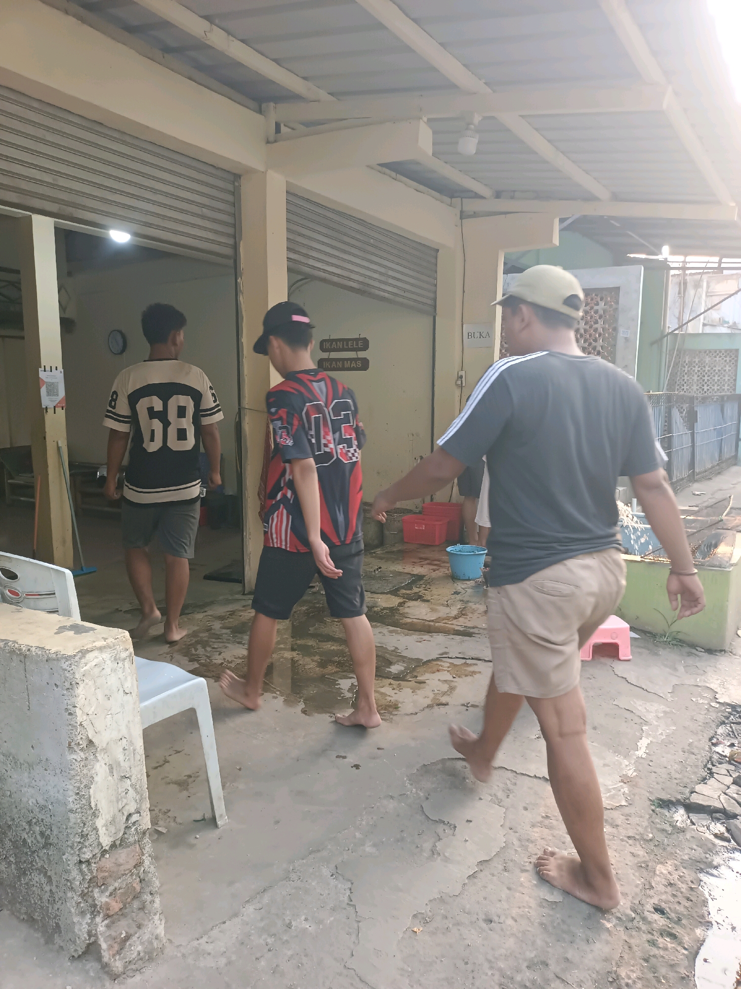 Three men are walking on a wet, concrete ground outside of a building with corrugated metal shutters and a metal roof. The man on the left wears a black and cream-colored jersey with the number "68" on the back and grey shorts. The middle man wears a black, red, and white patterned jersey with the number "03" on the back and black shorts. The man on the right wears a dark t-shirt with white stripes on the sleeves, tan shorts, and a light-colored baseball cap. He is holding out his left arm towards the middle man, and both appear to be walking away from the building. In the background, there are two red plastic bins, a blue bucket, and a pink plastic stool. Signs on the wall of the building read "IKAN LELE" and "IKAN MAS", which translate to "Catfish" and "Goldfish" respectively, suggesting this is a fish market or restaurant. A sign on the building also reads "BUKA", meaning "Open". The overall environment appears to be an outdoor area of a business in the city of Karawang, Indonesia, possibly in the morning or late afternoon given the sunlight.
