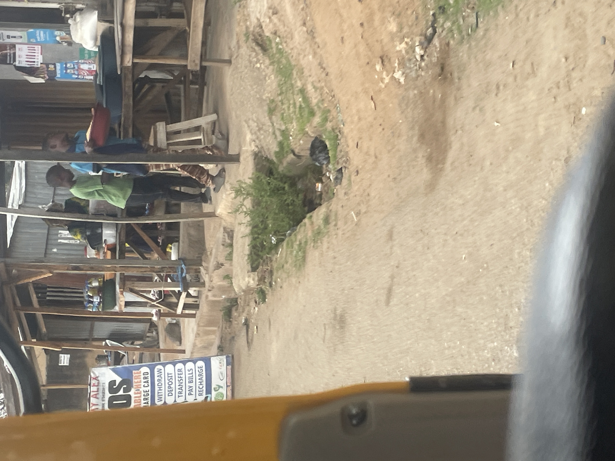 The image, taken from within a vehicle, shows a street scene in Jos, Nigeria. Visible is a dirt road with sparse vegetation along the side. A small commercial building, constructed of wood and corrugated metal, is situated to the left of the road. A sign on the building advertises financial services such as deposits, withdrawals, and bill payments. Several people are standing or walking near the building.