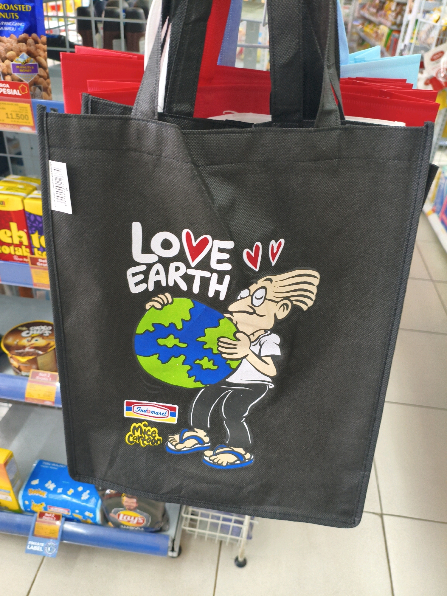 This is a close-up, eye-level, indoor shot of a black reusable tote bag with a cartoon image on it, displayed in a retail store. The bag features the text "LOVE EARTH" with a red heart symbol for the "O" in "LOVE" and two smaller red heart symbols. Below the text, a cartoon character with blond hair, glasses, and a white t-shirt is depicted holding a green and blue planet Earth. The character is wearing blue flip-flops. A logo that reads "Indomaret" with a yellow and blue emblem is visible on the lower left of the bag, followed by "Mice Cartoon" in yellow. The bag is one of several similar bags, with red and blue bags visible behind it. The background consists of store shelves stocked with various products, including packaged nuts, snacks, and other items. The floor is tiled. The retail environment suggests the location is a convenience store or supermarket. The presence of an "Indomaret" logo indicates the location is likely within an Indomaret store in the city of Sumberejo Kulon, Indonesia.