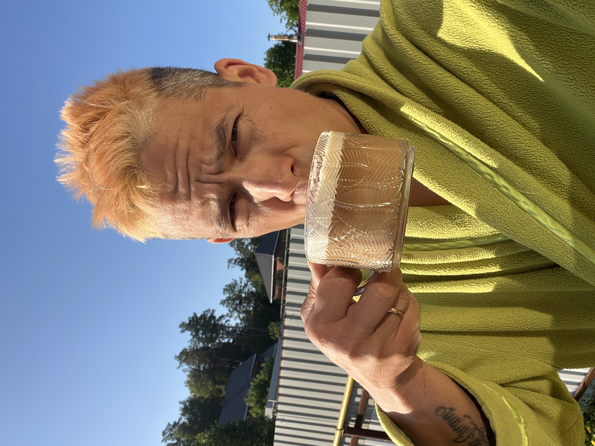 A man with dyed orange hair is wearing a light green fleece robe and holding a glass with a white, frothy beverage. The man has a tattoo on his forearm and a wedding ring on his finger. The background shows trees, houses, and a blue sky. The picture was taken outdoors, likely in the city of Chaikovsky, Russia.