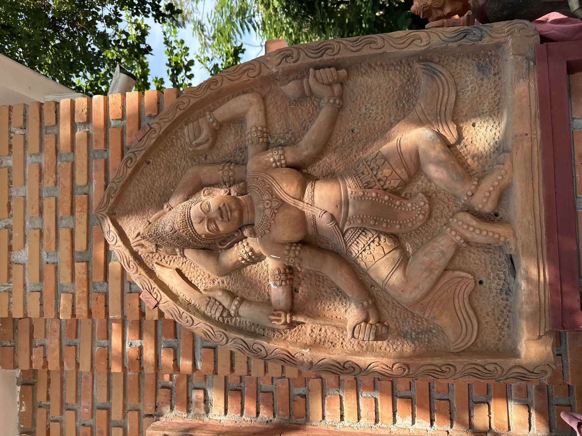 An elaborately carved terra cotta relief depicting a multi-armed deity stands against a brick wall in Nha Trang, Vietnam. The deity, possibly a Hindu or Buddhist representation, is adorned with intricate jewelry, a headdress, and patterned clothing. The figure has multiple arms, some holding symbolic objects such as a trident and a flame. The relief is set within an arched frame with decorative carvings. To the right, lush green trees and a bright blue sky are visible, suggesting an outdoor setting. A white security camera is mounted on the wall near the top right corner. The brick wall shows signs of weathering and age. The overall scene conveys a sense of ancient art and cultural heritage within a modern context.