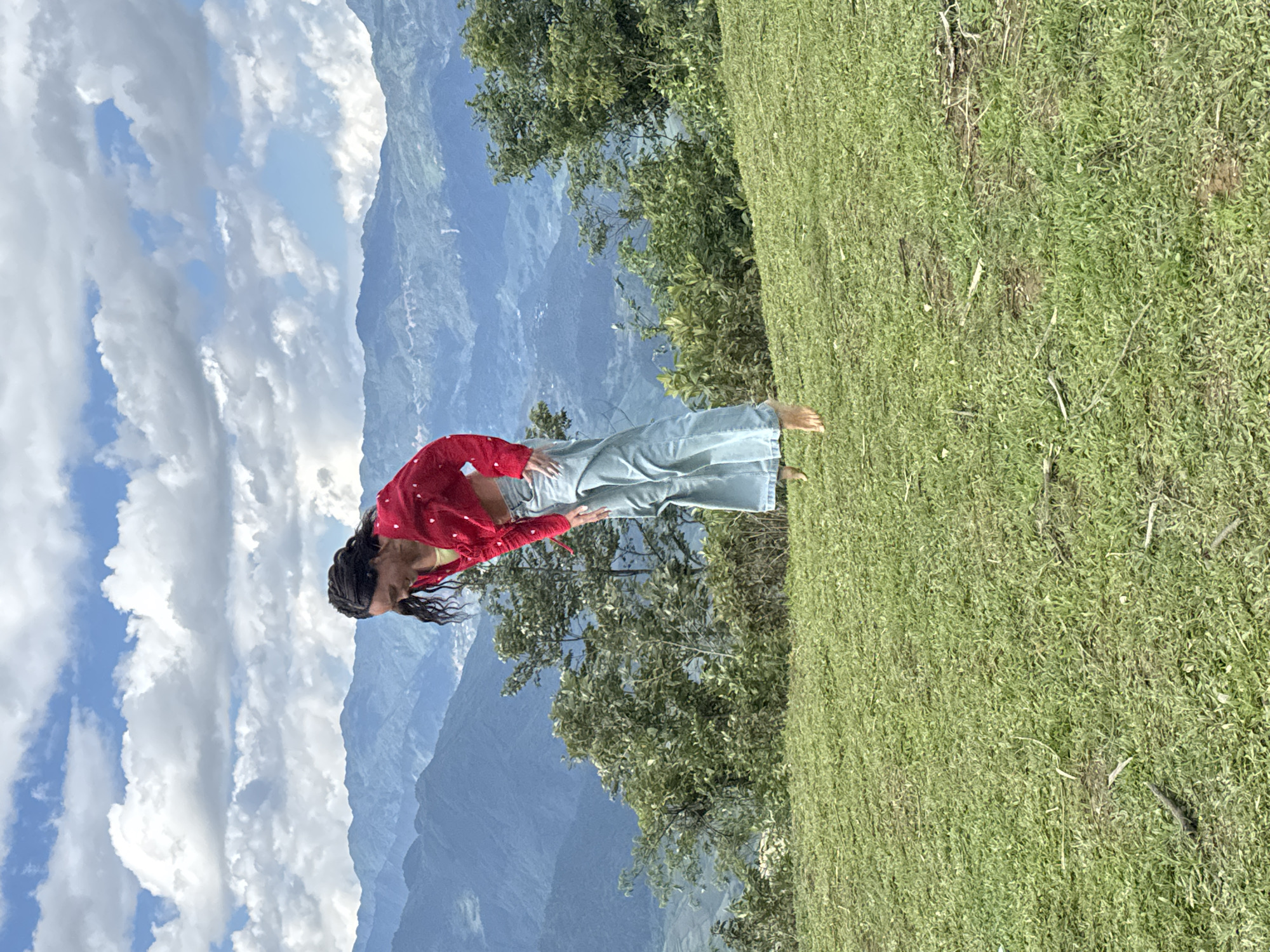 A vertical outdoor photograph taken in Tu Le, Vietnam, shows a single person captured mid-air, suspended several inches above a grassy field. The subject, appearing female, has dark braided hair and is dressed in a red long-sleeved top patterned with small white shapes and light blue wide-leg jeans, with bare feet visible. Her head is tilted downwards, and her expression suggests a slight smile or amusement. Her arms are bent, with hands positioned near her torso. The immediate foreground consists of a vibrant green grassy expanse, containing scattered dry patches and small twigs. A line of dense green trees and bushes forms the midground, separating the field from the distant background. The background features multiple layers of blue-tinted mountains extending across the horizon, with faint indications of distant settlements or structures. The sky above is bright blue, filled with numerous large, white cumulus clouds.