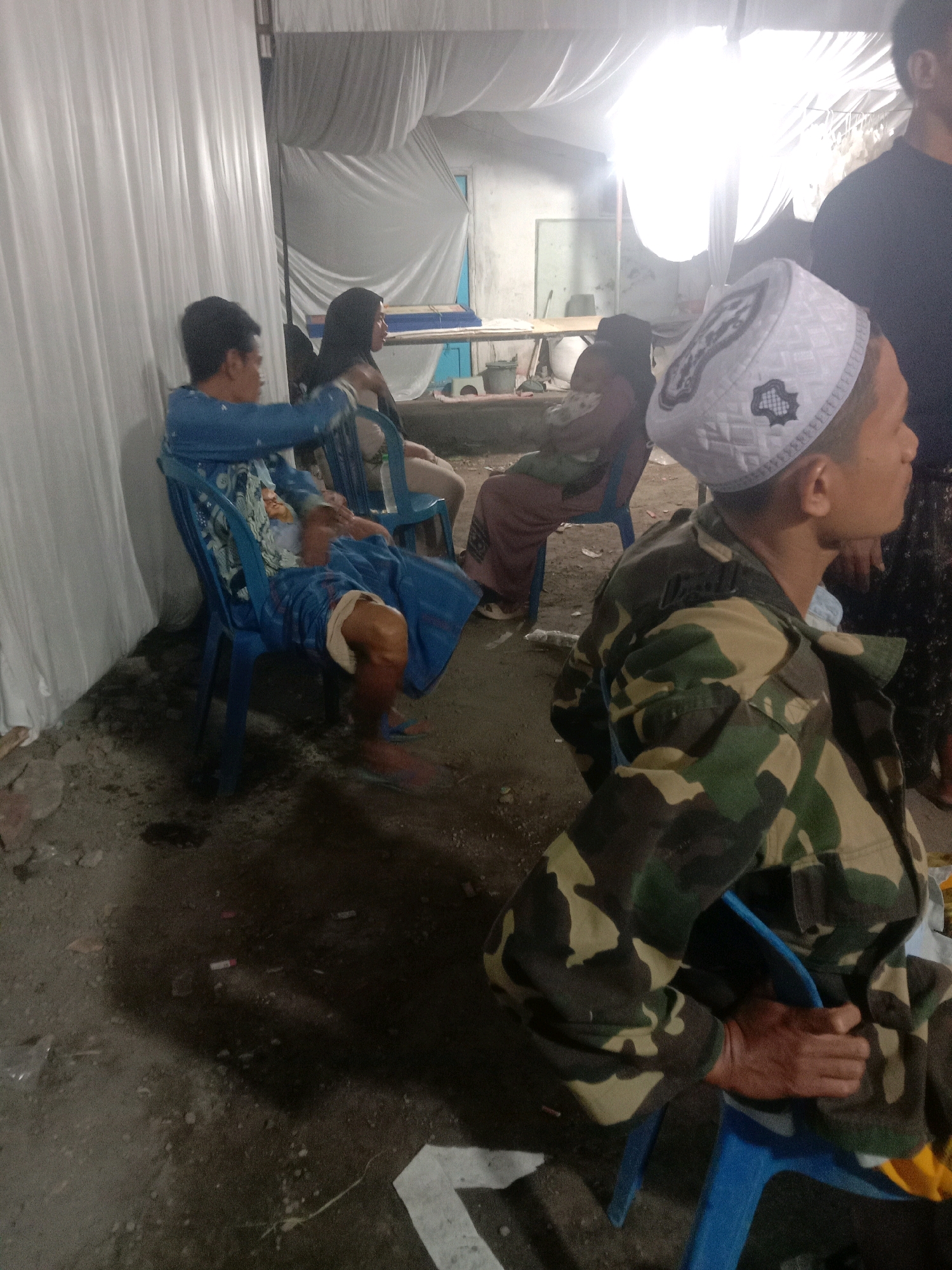 In the city of Karang Anyar, Indonesia, an indoor gathering is taking place, possibly a community event or a private ceremony, indicated by the white tent-like structure providing overhead cover. The scene is lit by an overhead light source, creating a bright, blown-out effect in the upper portion of the image.
In the foreground, a man wearing a camouflage jacket and a white peci (a cap worn by Muslim men) is seated on a blue plastic chair, facing towards the right of the frame. His right hand is resting on his lap. To his left, another man is seated on a similar blue chair, leaning back. He is wearing a blue patterned shirt and shorts, with his right leg bent and his foot resting on the floor.
Further back, two women are seated on chairs. One, on the left, is wearing a dark headscarf and a light-colored outfit. She is looking towards the right. The other woman, to her right, is also wearing a headscarf and a long, brown garment. She is seated behind a blue chair and is looking in the same direction as the woman next to her.
The environment appears to be an outdoor or semi-outdoor space that has been set up for an event. The ground is a mix of dirt and debris. Scattered on the ground are various items including cigarette butts, plastic wrappers, and a white piece of paper. The overall impression is of a functional, possibly makeshift, gathering space.