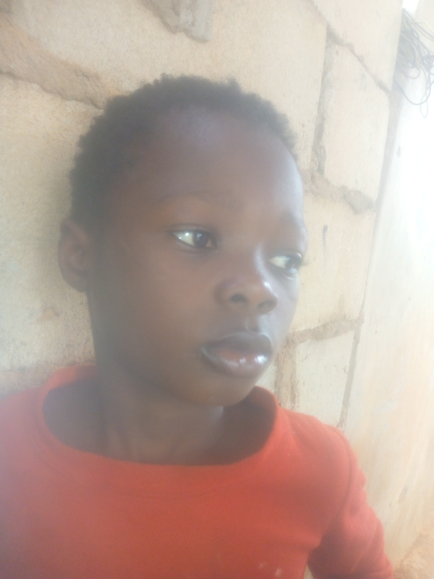A close-up, brightly lit portrait features a young person, likely a child, with dark skin and short, dark, natural hair. They are wearing an orange or reddish-orange long-sleeved top. The person is positioned in front of a light-colored wall constructed of bricks or blocks, with visible mortar lines and a rough texture.
The subject is looking towards their right (viewer's left) with a neutral, slightly contemplative expression; their lips are slightly parted. The lighting is very strong, causing some overexposure, particularly on the right side of the image and parts of the background, suggesting bright daylight. There is a sheen on the person's skin, especially on the forehead and lips. In the background, on the far right, dark wires or cables are visible against the wall. No text is visible in the image.