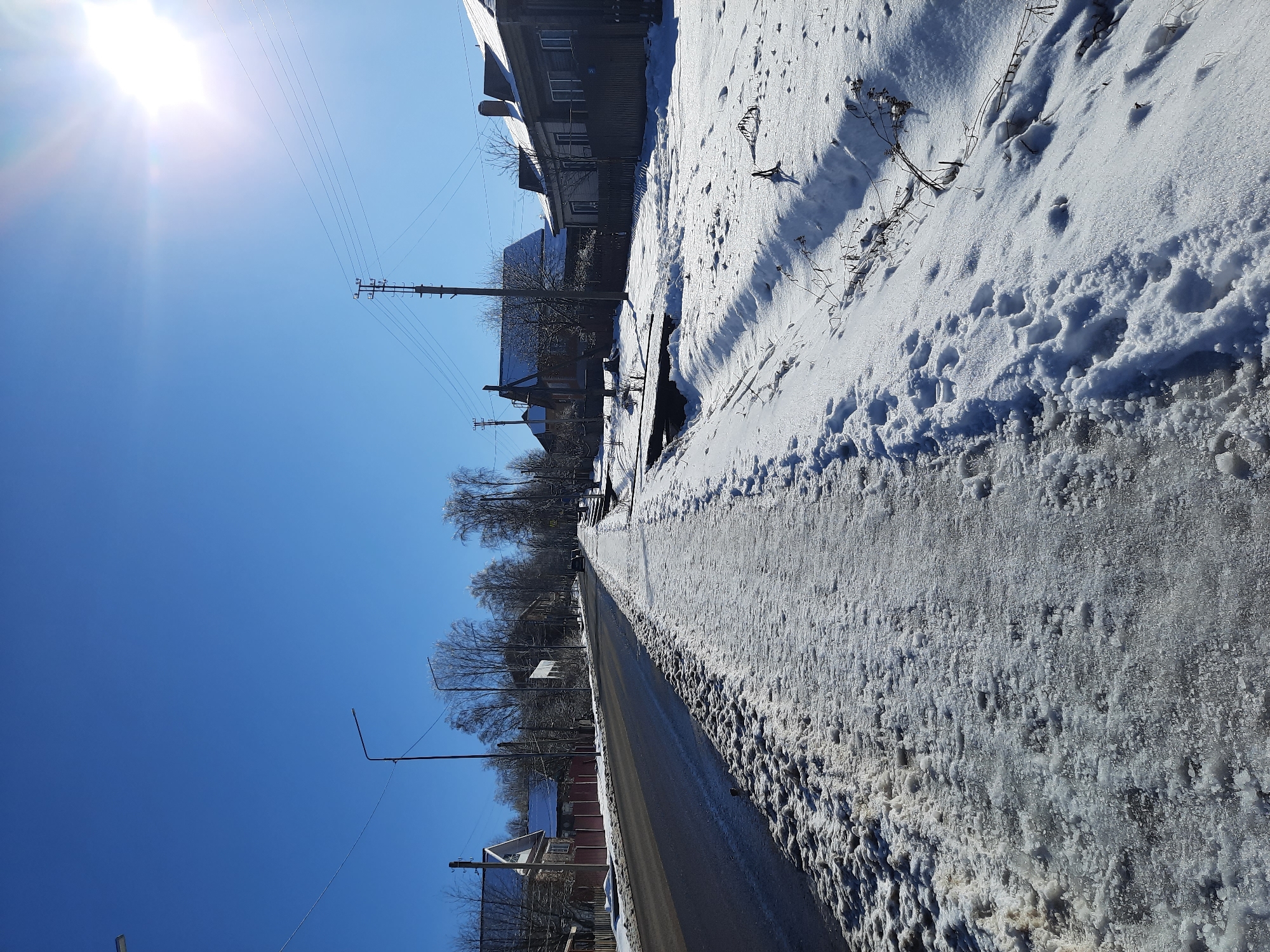 The image shows a winter scene in what appears to be a residential area in Krasnogorskoye, Russia. A road covered with snow and ice runs through the scene. One side of the road is bordered by a snow-covered sidewalk or shoulder, showing footprints. Houses are visible on both sides of the road, some partially obscured by trees. Power lines and utility poles stretch along the roadside. The sky is clear and blue, with the sun shining brightly. The image is slightly rotated.