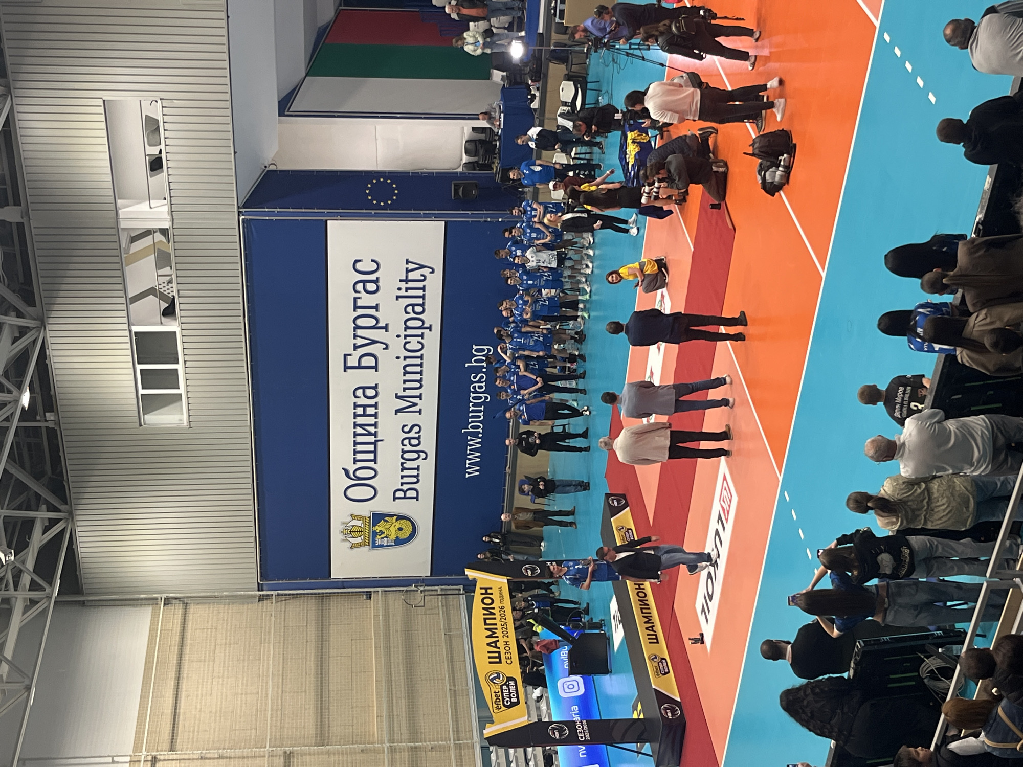 The image depicts a group of people gathered on a two-toned blue and red surface with white lines, likely a volleyball court. A large banner in the background, featuring a blue and white design, displays the text "Община Бургас Burgas Municipality" and "www.burgas.bg". The banner also includes a seal, indicating that the location is in the city of Burgas, Bulgaria.
The people are standing in rows, suggesting a team or organized group. Some are in blue sports uniforms, while others are in casual attire. Some individuals are taking photographs, and there are additional banners with the text "ШАМПИОН", "Efbet СУПЕР ВОЛЕЙ", and "СЕЗОН 2025/2026 ГОДИНА". There are also other logos, including the "efbet", and "nvBu" logo and the Instagram logo. A person, presumably "Дарин Миров", wears the number "2" on his back. The area appears to be an indoor sports venue with visible roofing and beams.