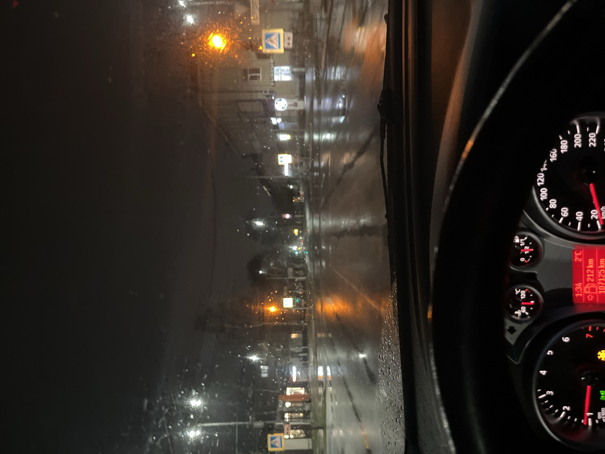 The image depicts a view from inside a car driving along a wet street at night in what appears to be the city of Chaikovsky, Russia. Streetlights illuminate the road, reflecting on the wet surface. Building facades and pedestrian crossings are visible along the sides of the street. The car's dashboard is partially visible, indicating the time as 1:34, an outside temperature of 2 degrees Celsius, a distance of 212 km, and a total odometer reading of 107125 km. The speedometer needle is close to zero, indicating the car is either stopped or moving very slowly. The sky is dark and the overall light condition is low.