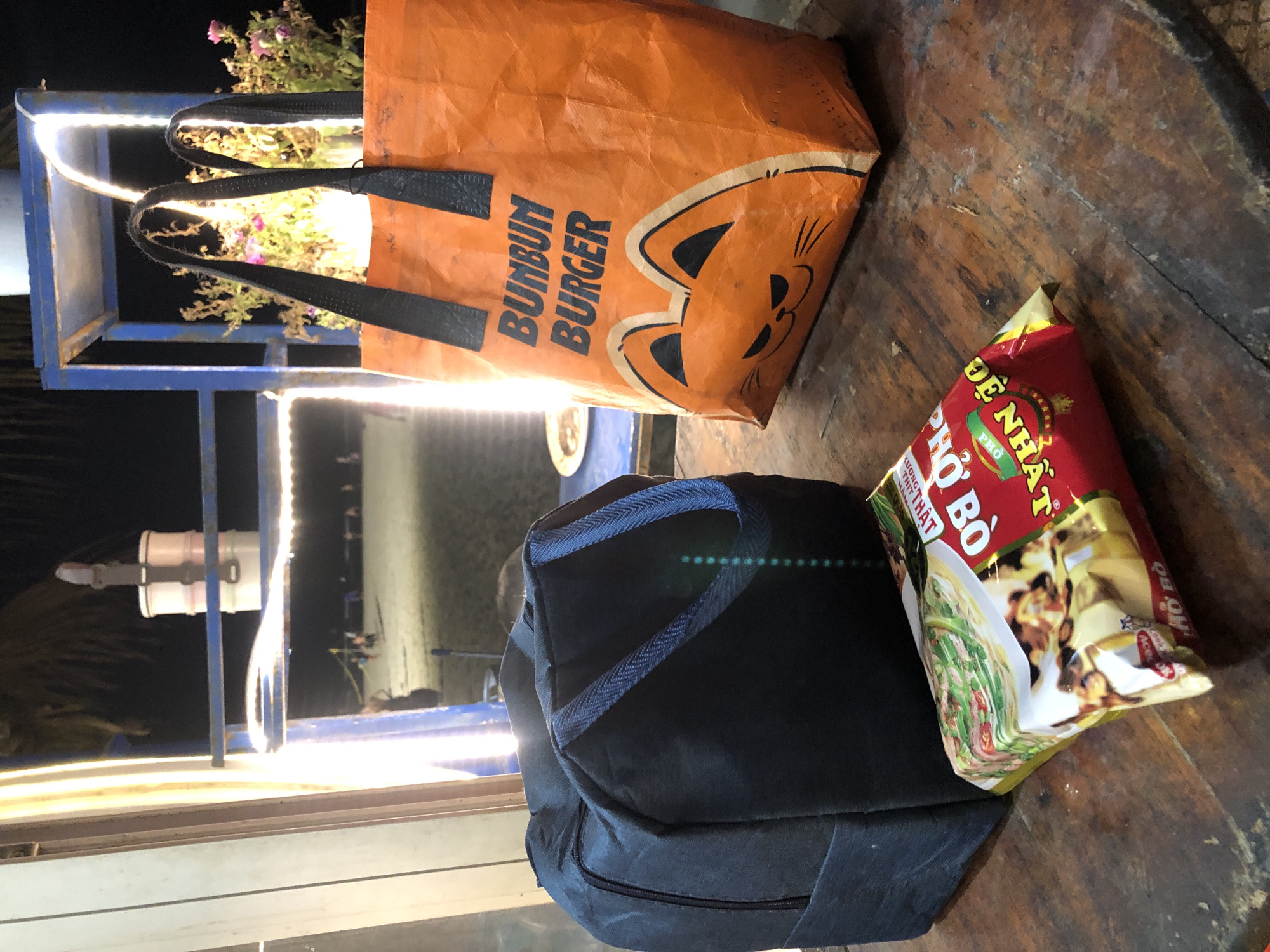 The image shows an assortment of items on a rustic wooden table, suggesting a casual outdoor setting, possibly near a food stall or market given the presence of food packaging and bags. In the foreground, a dark blue fabric bag with blue zipper detail is prominently displayed. To its right, a red and yellow packet of instant noodles, "PHO BO," is visible. The text "PHO BO" is clearly legible, and partially visible text includes "DE NHAT" and "XUONG," indicating a Vietnamese product.
Behind the blue bag, an orange reusable shopping bag with a cartoon cat face and the text "BUNBUN BURGER" is seen. The "BUNBUN BURGER" text is in a dark, bold font.
In the background, a blue metal structure with bright white LED lights is illuminated, casting a glow on a bunch of purple and pink flowers contained within the structure. The lighting suggests it might be evening or night. The overall scene appears to be set in a location with a warm, humid climate, consistent with Đà Nẵng, Vietnam. There are no people visible in the image, and the focus is on the objects and the immediate surroundings. The arrangement of items implies preparation for a meal or a recent purchase.