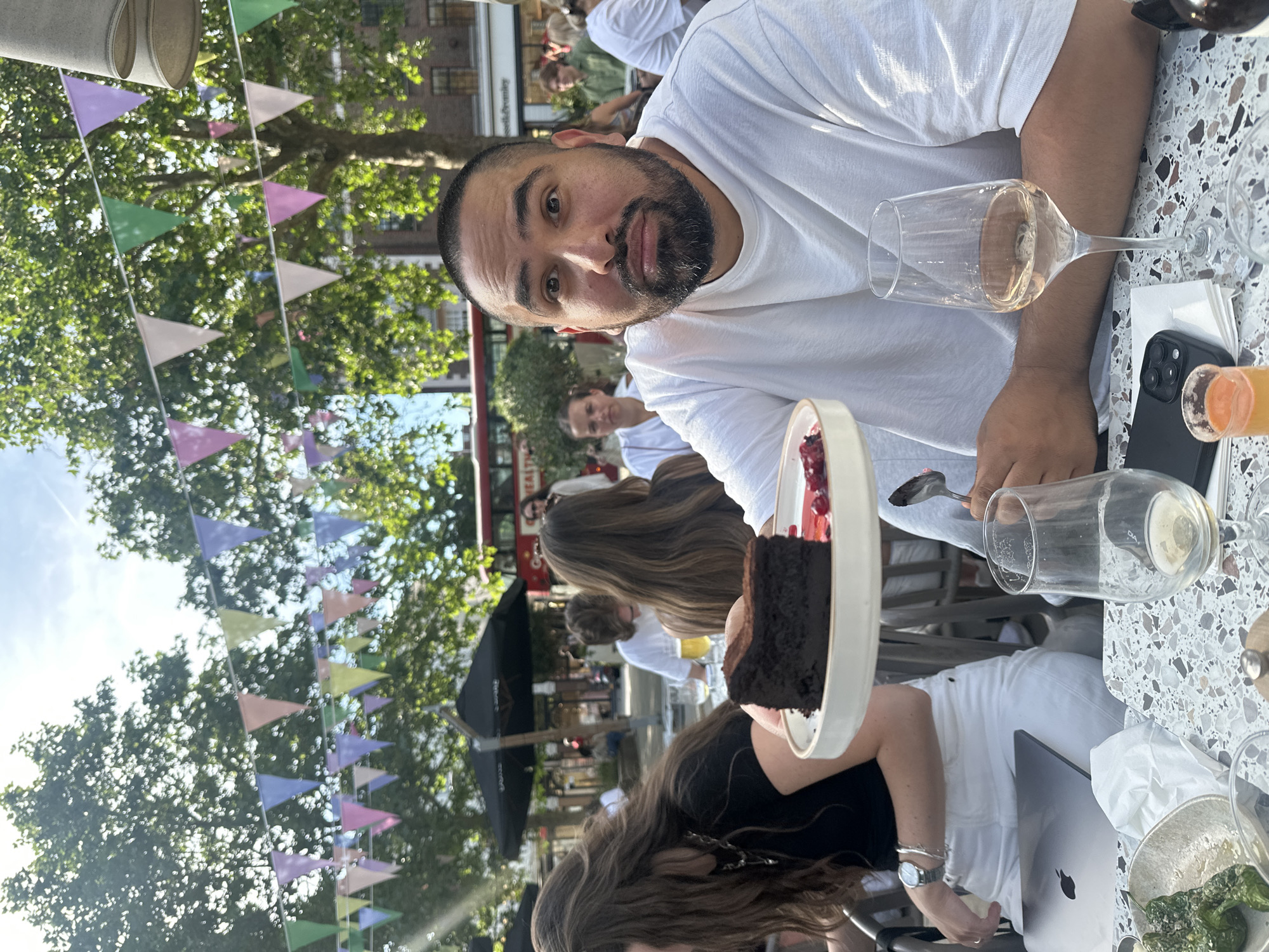 The image depicts a bearded man wearing a white t-shirt, seated at a table. He is looking at the camera with a slightly surprised expression. On the table is a glass of white wine, a smartphone, a glass with orange liquid, and a plate of chocolate cake.
Around him are several people, some seated at tables. There are colorful triangular flags strung overhead, and trees in the background. A double-decker bus and brick buildings are visible. The setting appears to be an outdoor dining area in the city of London, United Kingdom.
