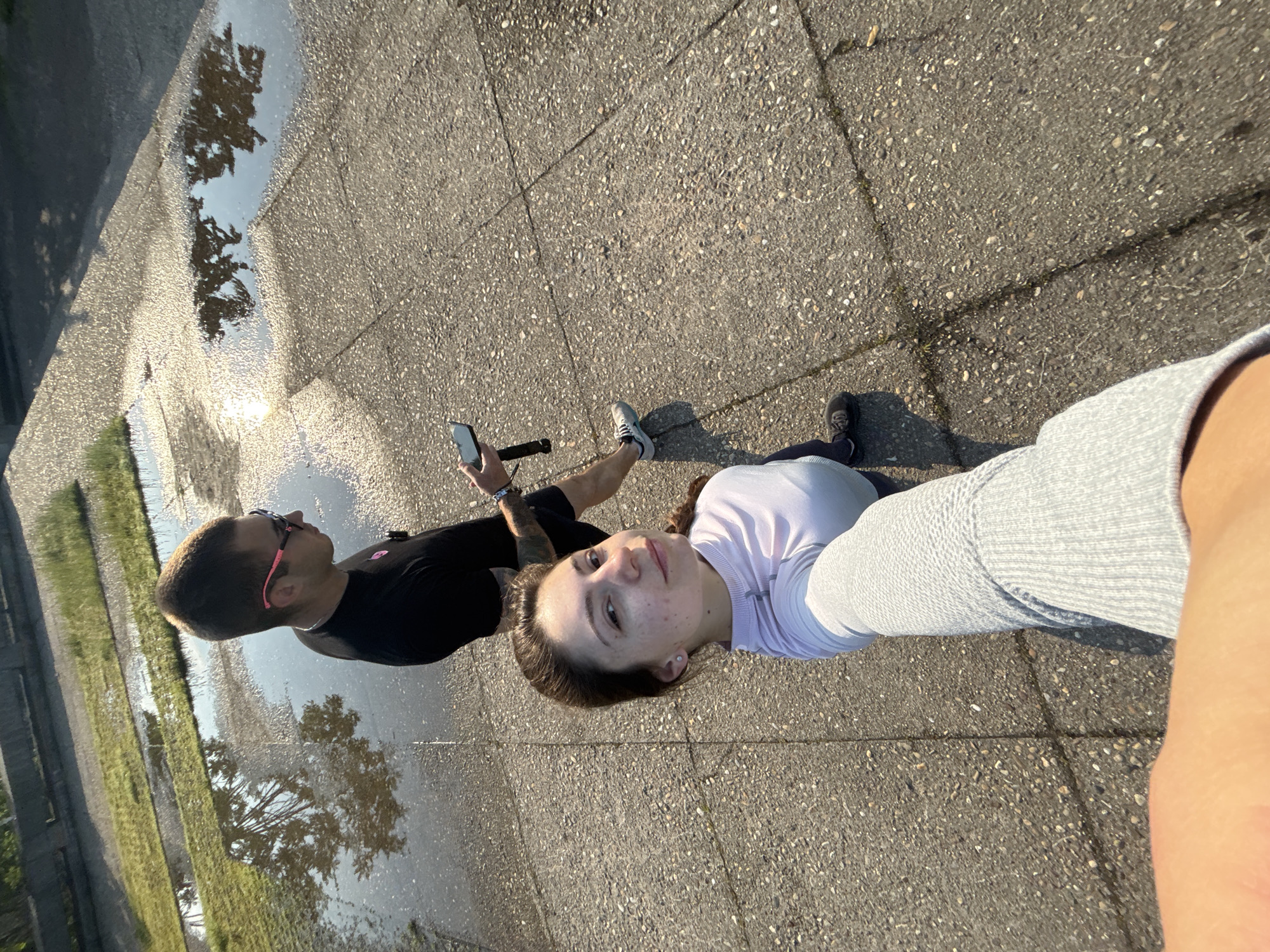 This media file captures a high-angle, rotated view of two individuals outdoors in Varna, Bulgaria. The primary subject, a young woman, is in the foreground, facing the camera with a neutral expression, suggesting she is taking a selfie. She has brown hair, fair skin with some freckles, and is wearing a light purple or white long-sleeved top and grey leggings. Her arm, holding the camera, is visible on the right side of the frame.
Behind her and slightly to the left stands a man with short brown hair, wearing a black t-shirt and sunglasses with distinctive pink frames. He has tattoos on his left arm and is holding a black smartphone, possibly attached to a stick or tripod. He is looking away from the camera, towards the left of the frame.
They are standing on a paved surface, likely concrete or asphalt, characterized by small, embedded pebbles and visible cracks. A long, narrow puddle stretches along the ground from the left side of the frame towards the background, reflecting the bright sky, a shining sun, and the silhouettes of trees, indicating a sunny day, possibly morning or late afternoon due to the strong sun reflection. In the distant background, patches of green grass and a grey structure are vaguely visible.
The overall scene suggests a leisurely outdoor activity, perhaps a walk or exploration. There is no visible text in the image.