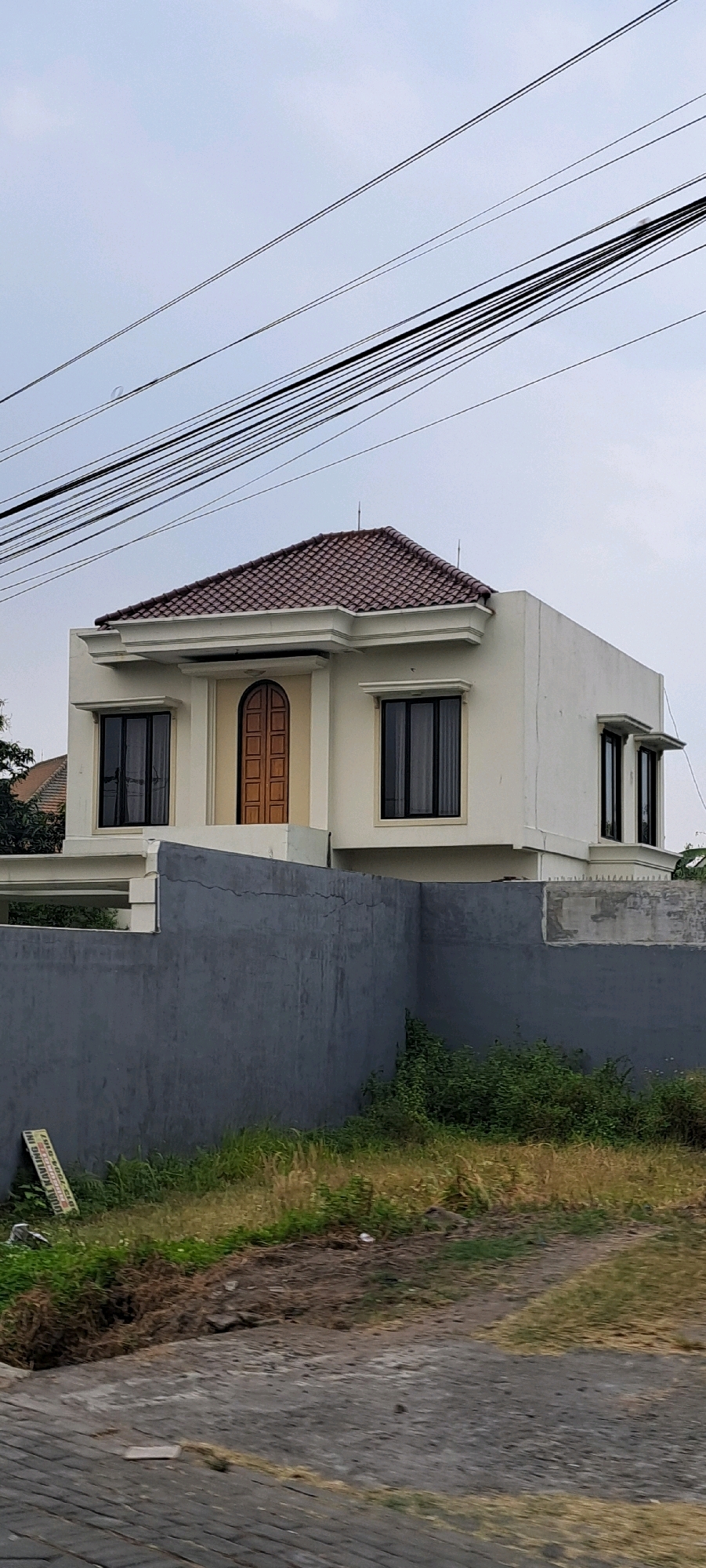 A contemporary, two-story house with a cream-colored facade and a tiled, reddish-brown roof is visible in Durensewu, Indonesia. The house features dark-framed windows and a prominent, arched wooden door on the front. A dark gray, textured wall encloses the property, with overgrown grass and weeds growing along its base. Overhead, a complex network of black electrical wires crisscrosses the pale sky. The foreground shows a paved area with a cobblestone pattern. The overall impression is of a well-maintained, modern residential property situated in a tropical urban environment.